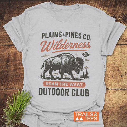 Buffalo Wilderness Club T-Shirt featuring a bison graphic with PLAINS & PINES Co. and ROAM THE WEST text, on a wooden surface with a pine sprig.