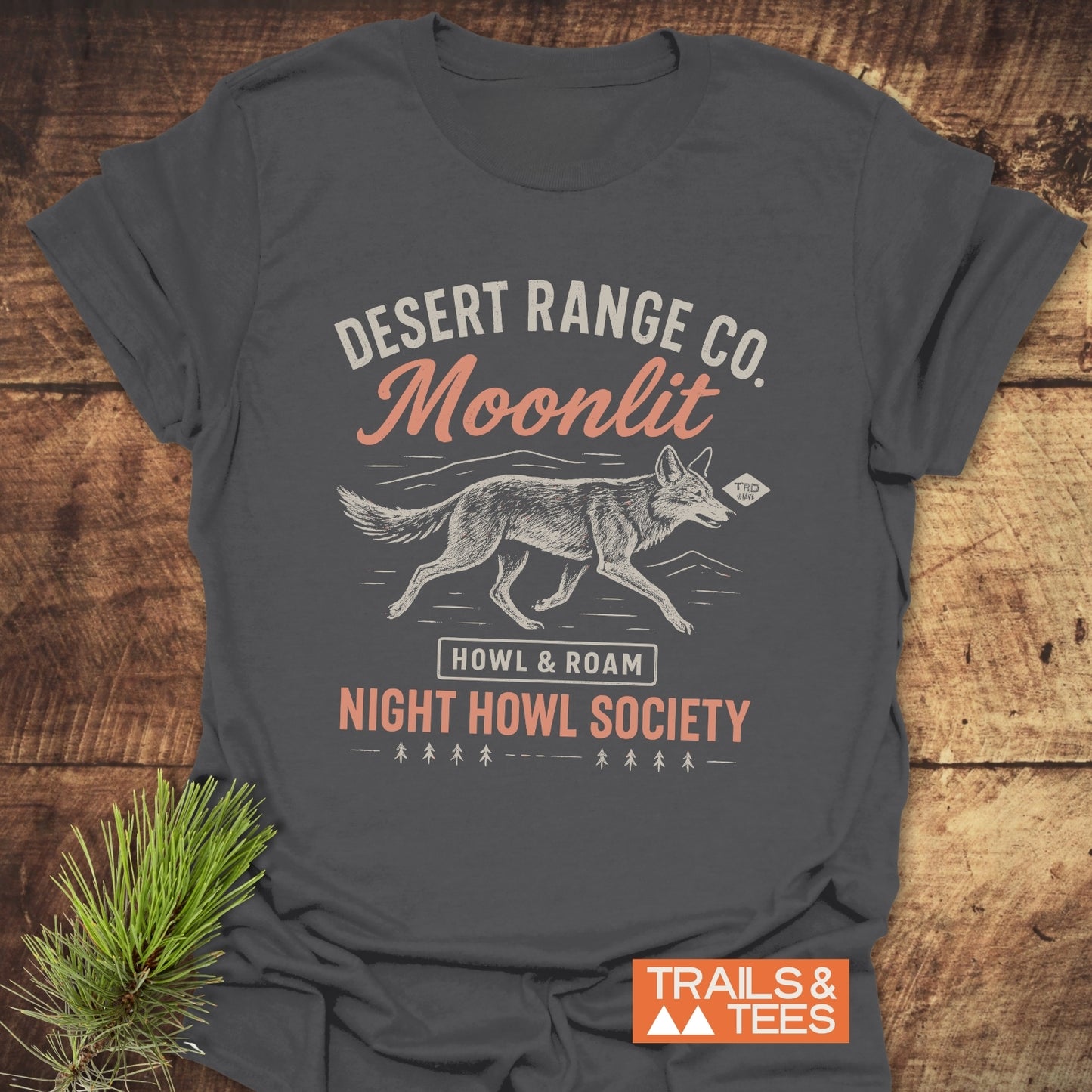 Night Howl Society T-Shirt featuring a stylized coyote print on charcoal-gray, made from soft ring-spun cotton. Laid on wood with an orange Trails & Tees logo.