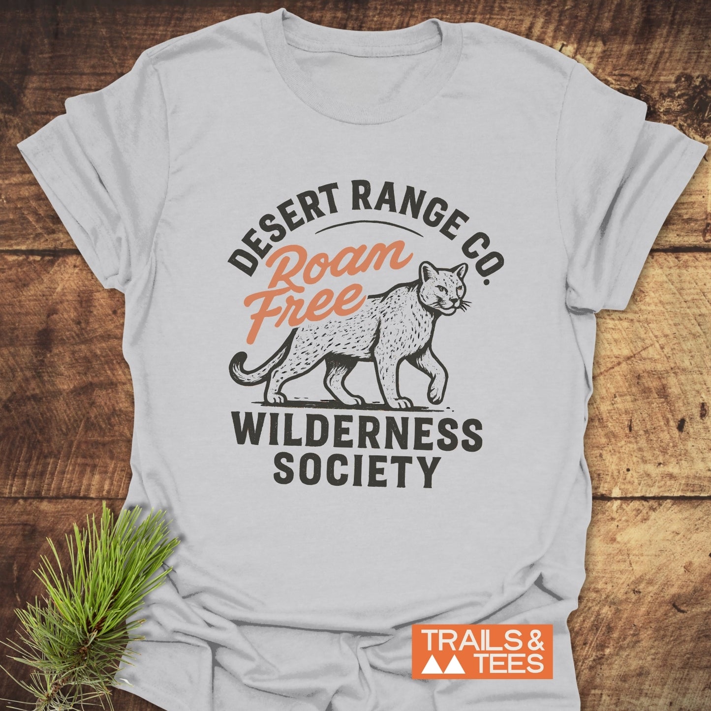 Roam Free Cougar T-Shirt featuring a mountain lion graphic and text, laid flat on wood, with pine sprig nearby; embodies comfort and outdoor adventure spirit.