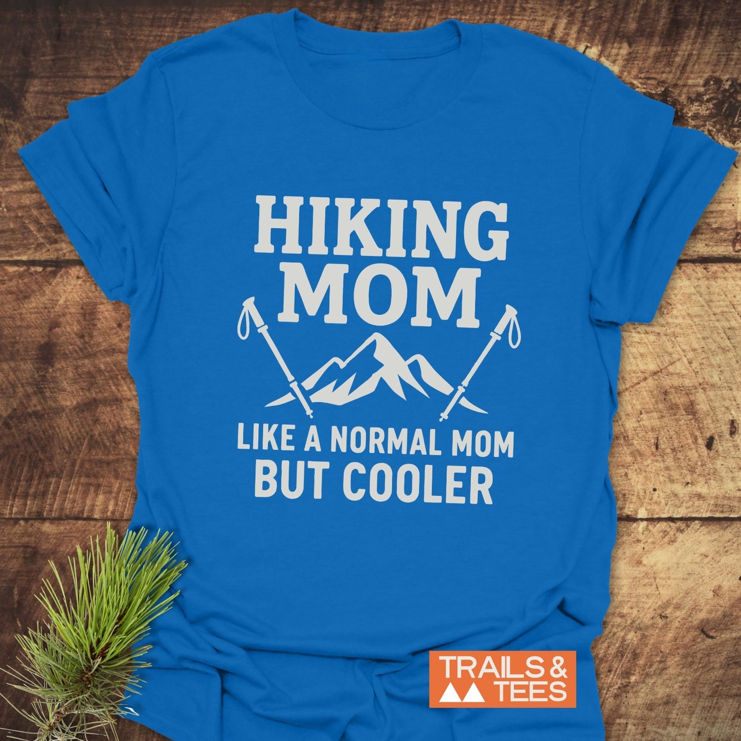 Hiking Mom T-Shirt with mountain graphic and hiking poles, on wood surface. Made from soft cotton for outdoor comfort, featuring durable print and pre-shrunk fit.