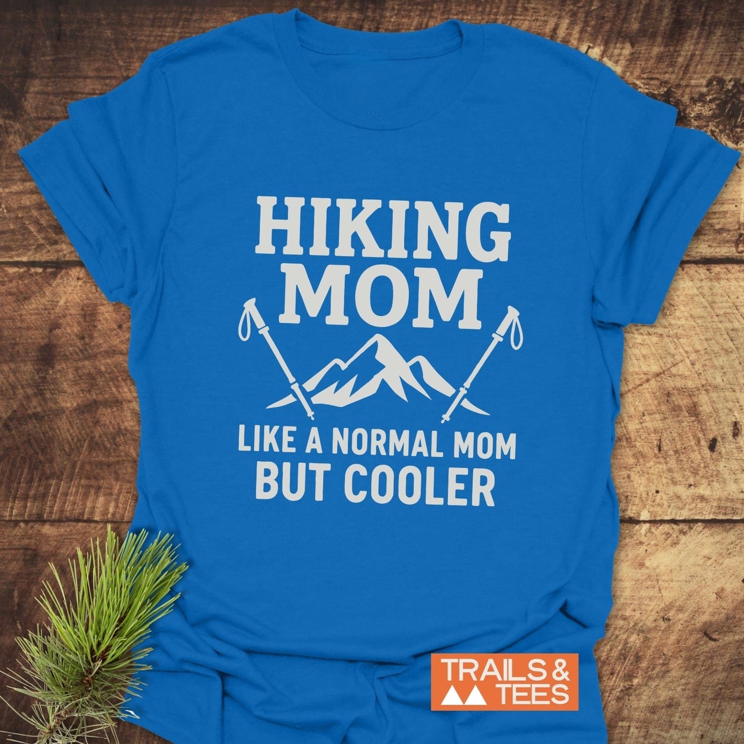 Hiking Mom T-Shirt with mountain graphic and hiking poles, on wood surface. Made from soft cotton for outdoor comfort, featuring durable print and pre-shrunk fit.