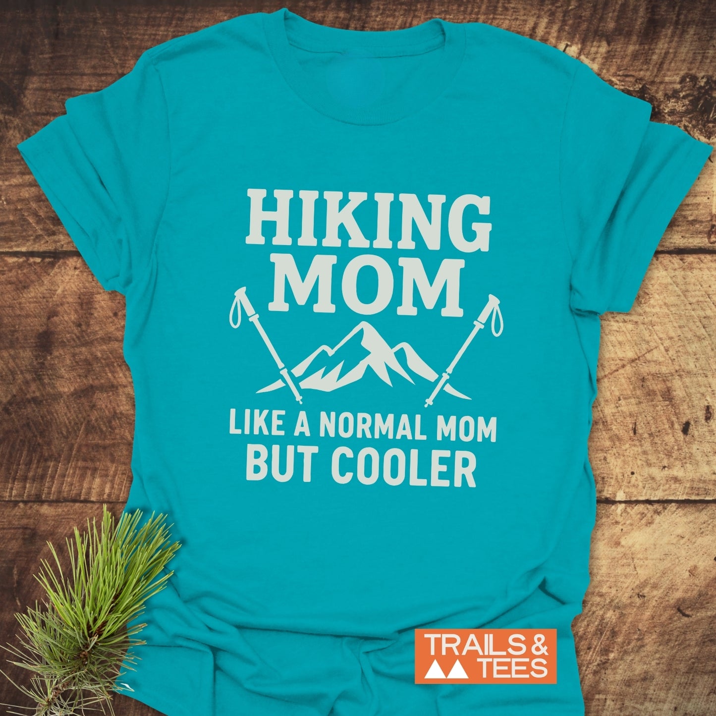 Hiking Mom T-Shirt with white “Hiking Mom” slogan and mountain graphic, laid on wood. Made from soft, durable cotton for outdoor enthusiasts.