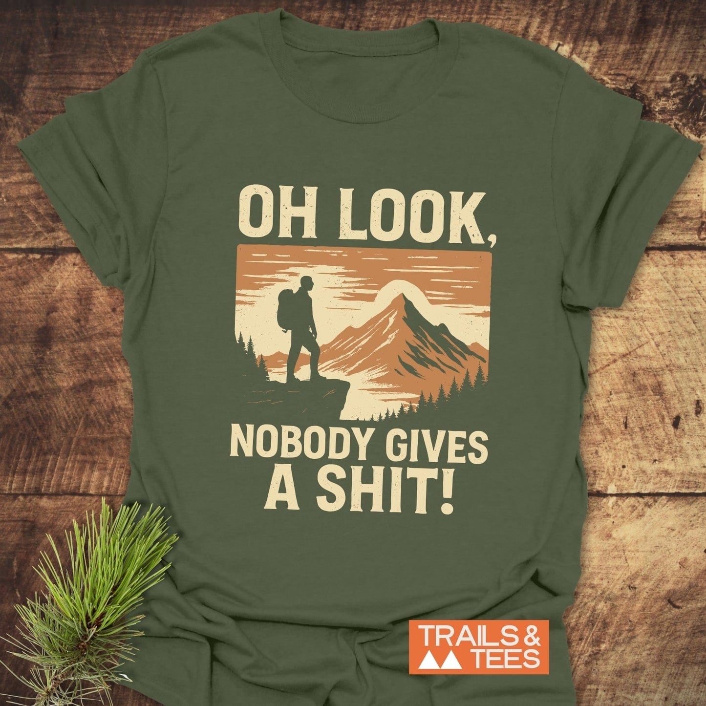 Olive-green Oh Look! Retro T-Shirt with a hiker graphic, bold text, and Trails & Tees logo, styled with a pine sprig on wood.
