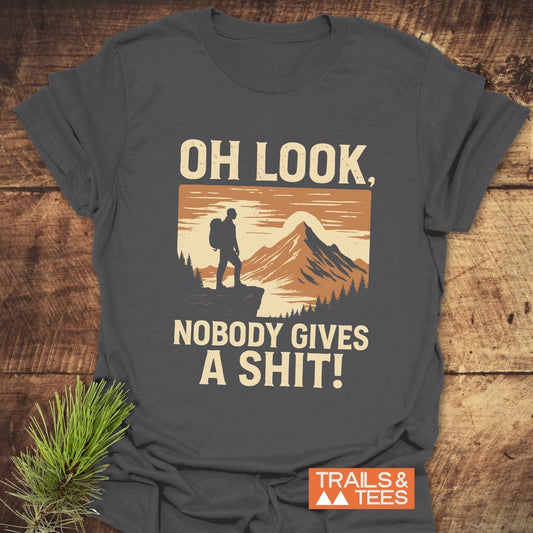 Oh Look! Retro T-Shirt featuring a hiker graphic, bold text, and Trails & Tees logo, laid on wood with a pine branch beside it.