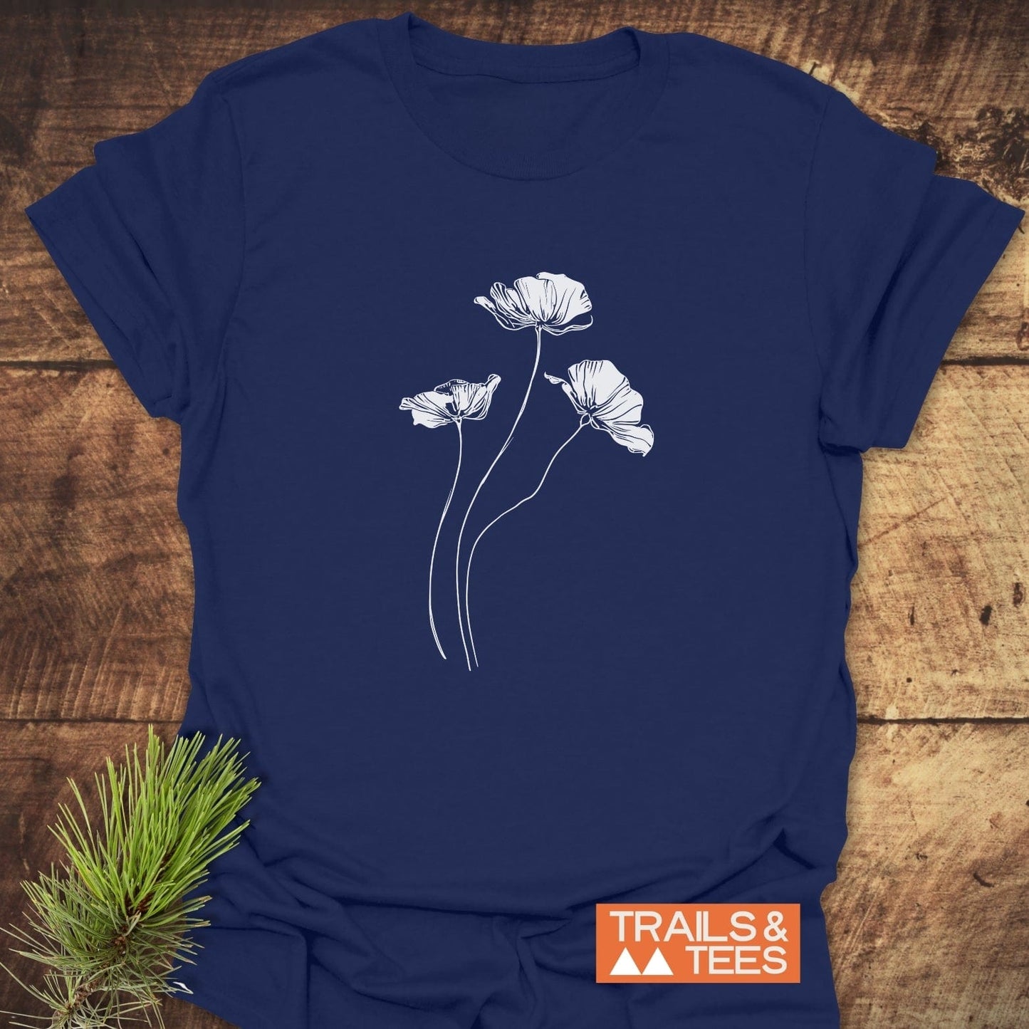 Delicate Poppies T-Shirt: Navy-blue tee with white poppy line-drawing, laid on wood surface, styled with pine needles, featuring durable, comfort-focused design.