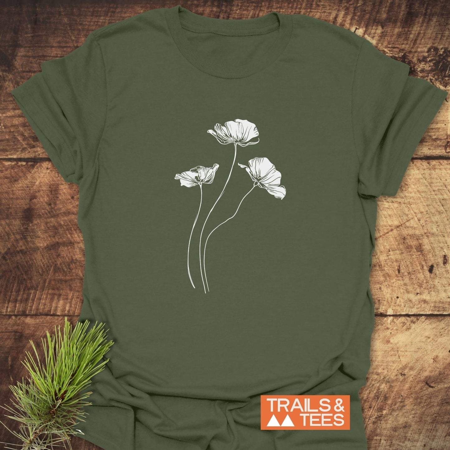 Delicate Poppies T-Shirt featuring white poppy drawings on olive-green fabric, with TRAILS & TEES logo, laid on rustic wood, accented by evergreen sprig.