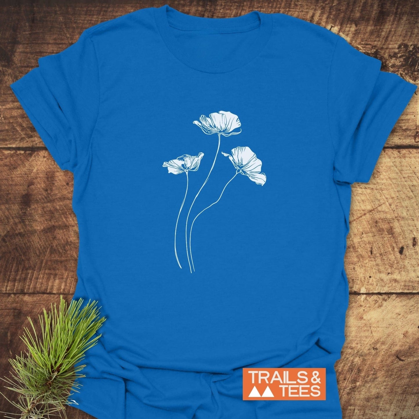 Delicate Poppies T-Shirt with white floral line-drawing on cobalt-blue fabric, featuring a “TRAILS & TEES” logo; designed for comfort and outdoor adventures.