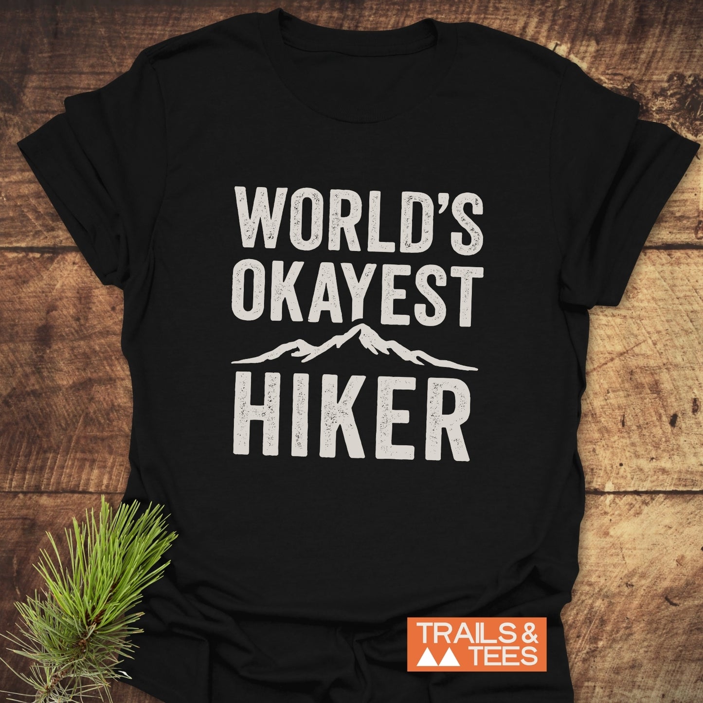 Black T-shirt with WORLD'S OKAYEST HIKER and mountain graphic, styled with pine sprig. Features durable print, ring-spun cotton for comfort, by Trails & Tees.