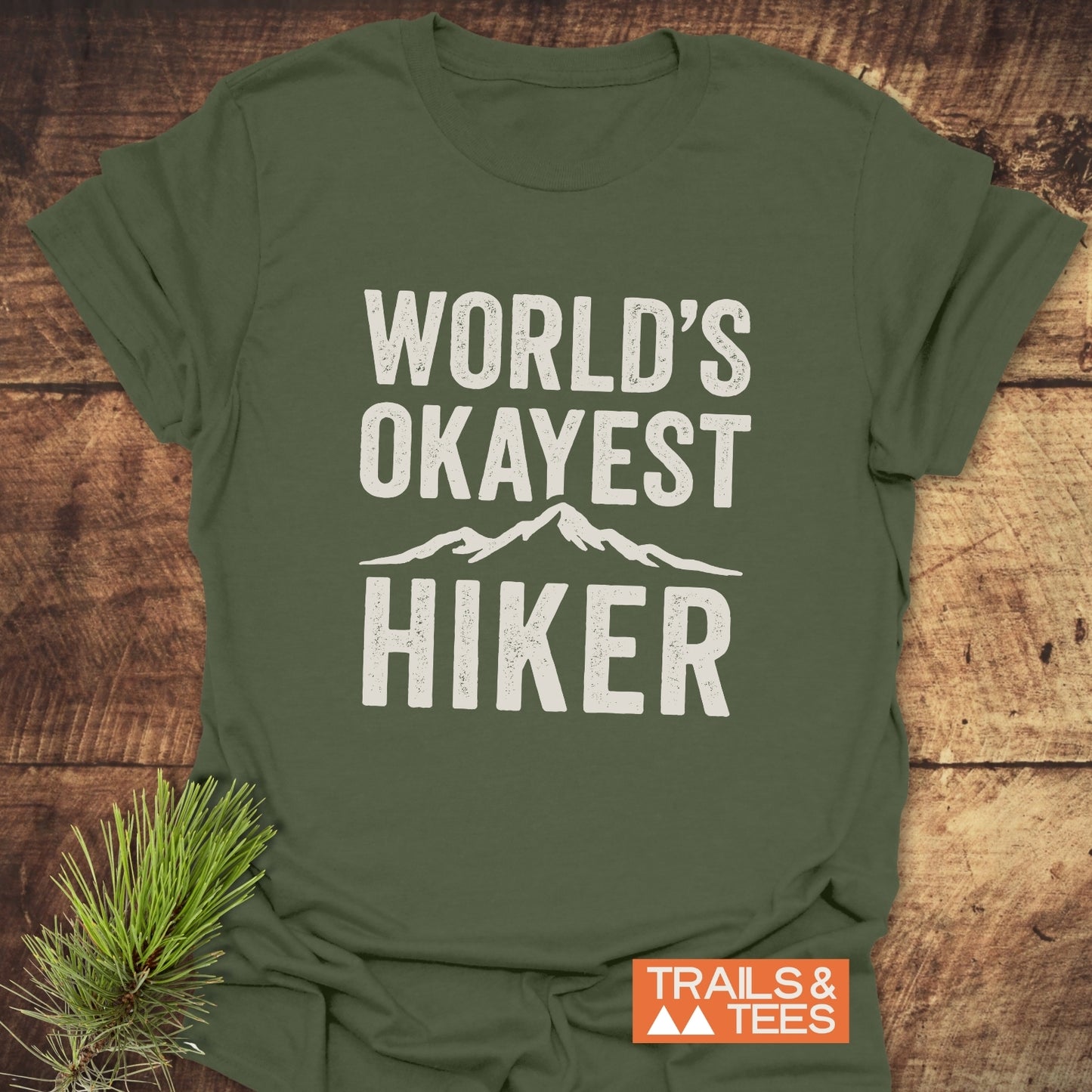 Forest-green World's Okayest Hiker T-shirt with distressed white text and mountain graphic, laid flat on wood, featuring Trails & Tees logo and pine needles.