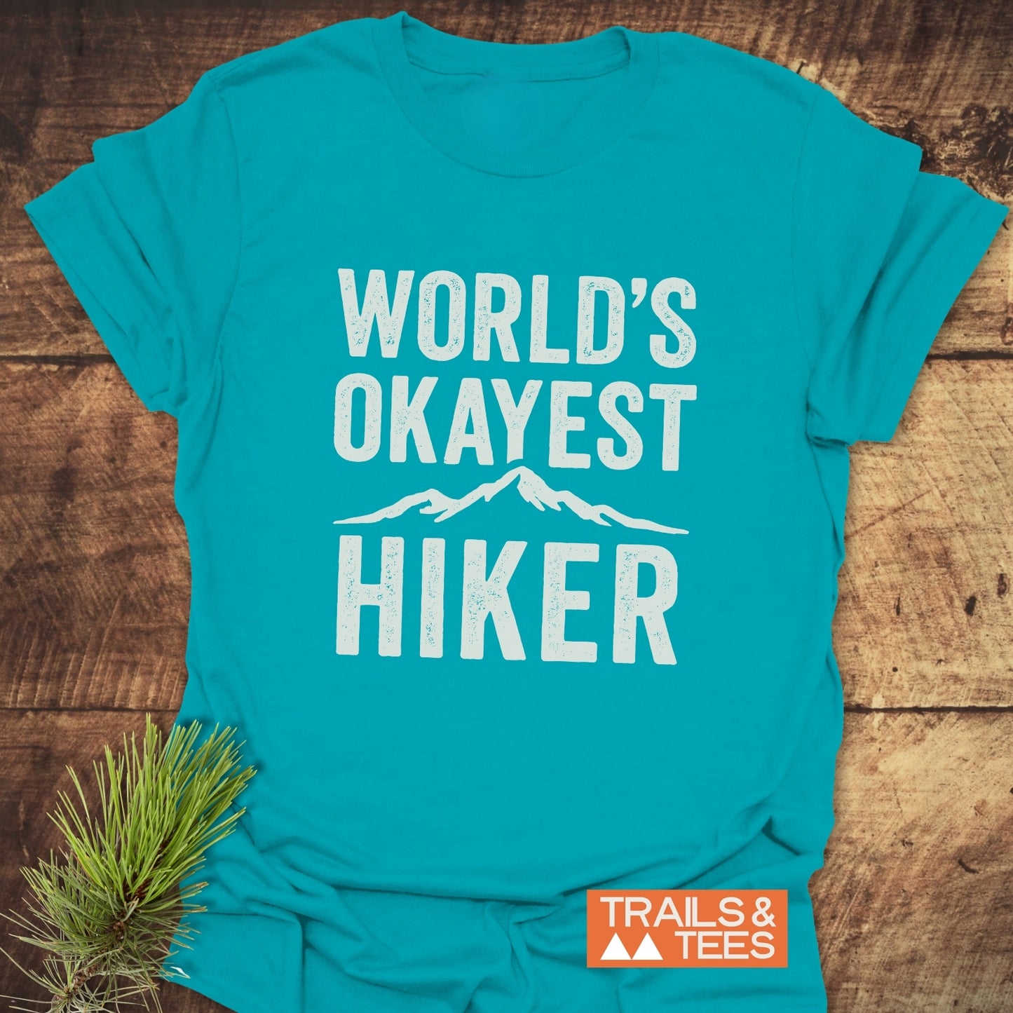 World's Okayest Hiker T-Shirt featuring a white slogan and mountain silhouette, with a Trails & Tees logo, laid on a wooden surface.