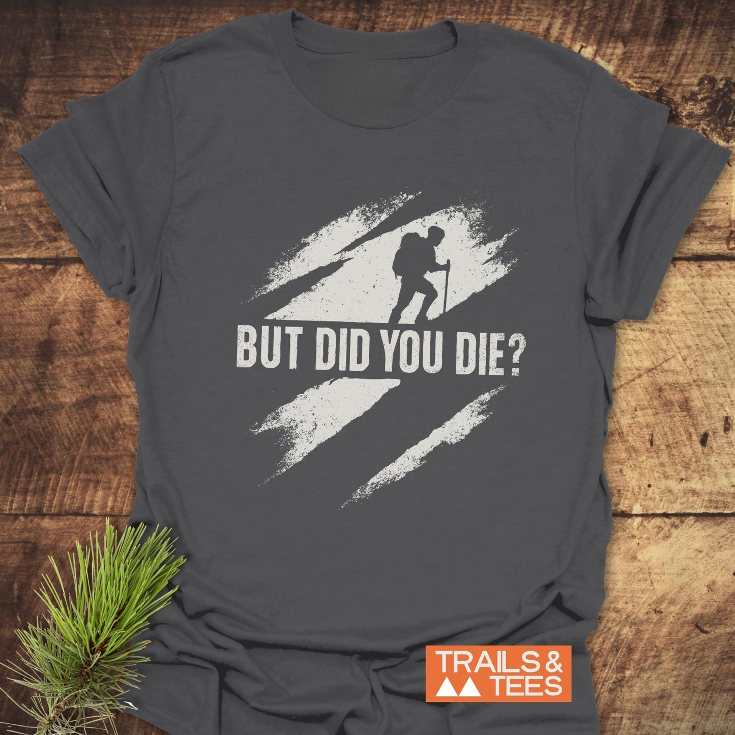 But Did You Die? Hiking T-Shirt: Charcoal-gray tee with a distressed backpacker graphic, 'TRAILS & TEES' logo, and pine sprig accent on wooden surface.