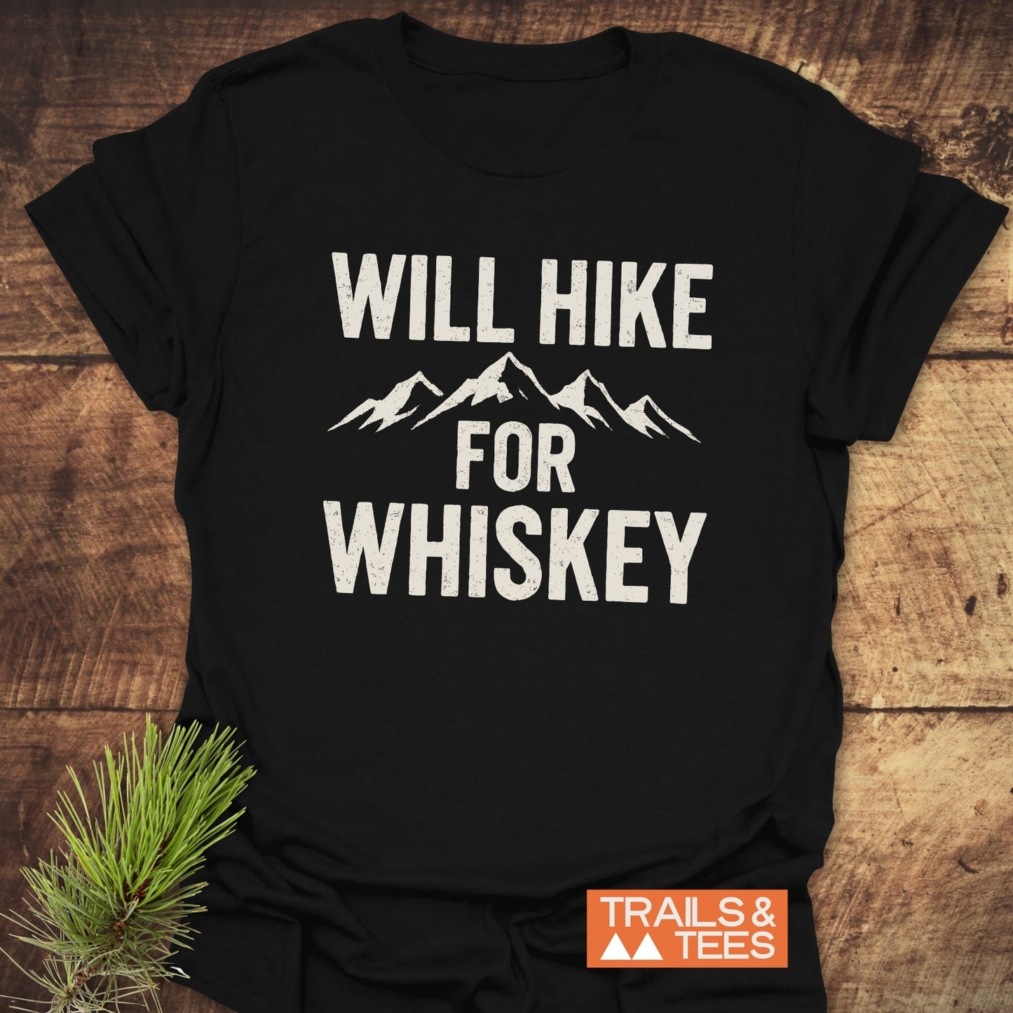 Will Hike For Whiskey T-Shirt with mountain graphic and slogan, laid on wood, featuring durable, lightweight cotton for outdoor enthusiasts. Trail-tested, pre-shrunk, eco-friendly design.
