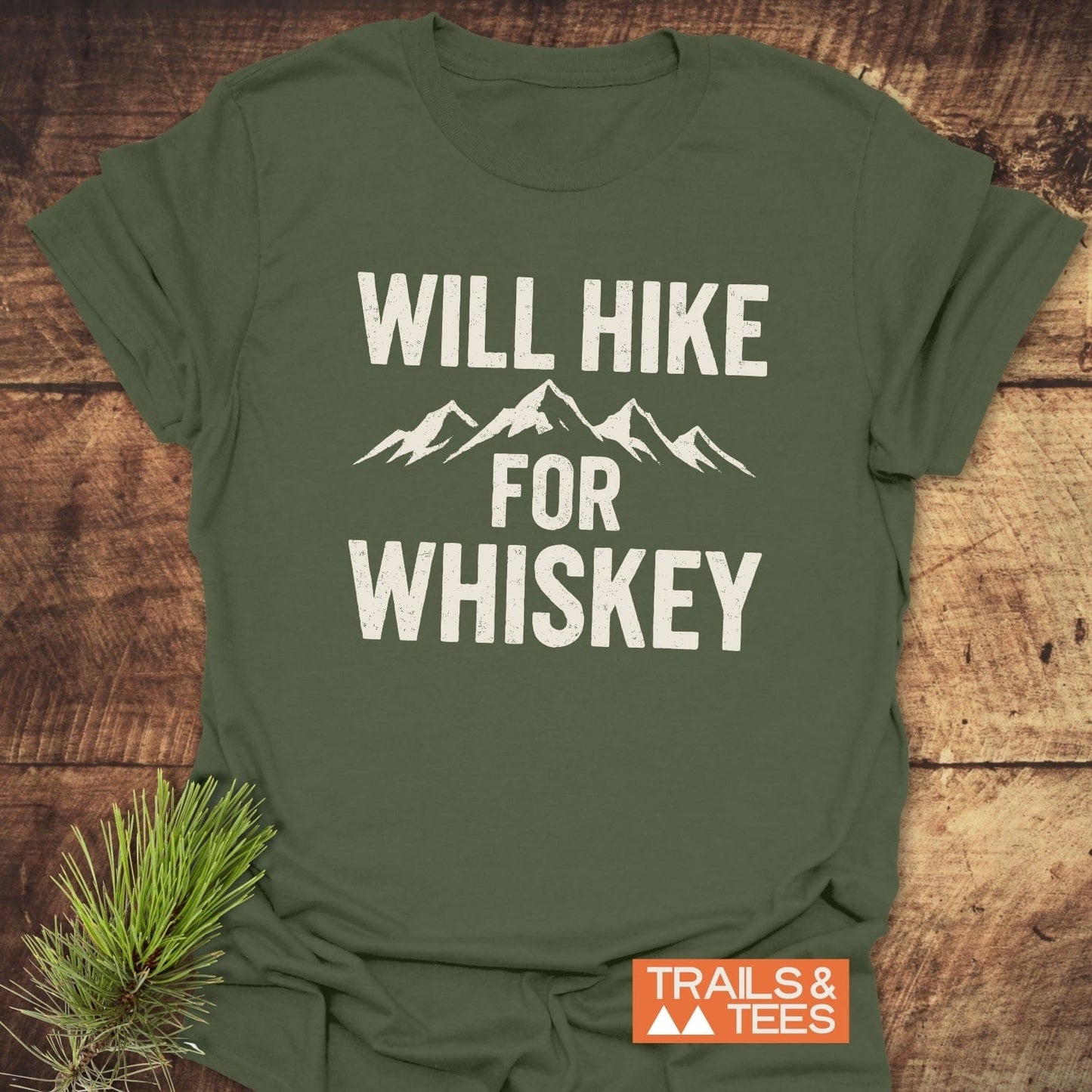 Will Hike For Whiskey T-Shirt with mountain graphic, displayed on wood, showcasing Trails & Tees label and pine sprig. Made from soft, lightweight cotton.