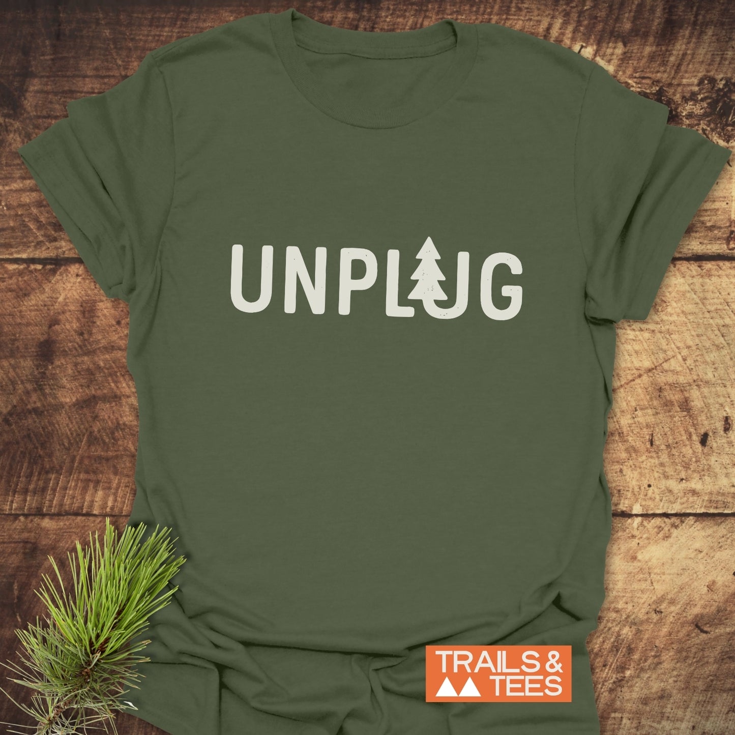 Unplug T-Shirt with a pine-tree design on olive-green fabric, showcasing lightweight, soft ring-spun cotton. Includes TRAILS & TEES logo and pine needle accent.