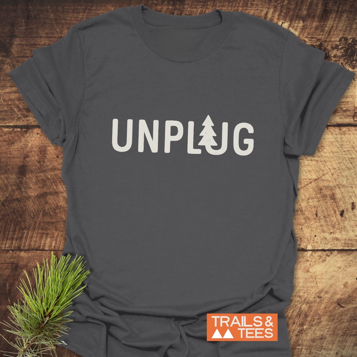 Unplug T-Shirt featuring UNPLUG with a pine-tree L on dark-gray cotton, laid on wood, accented by a pine branch and Trails & Tees logo.