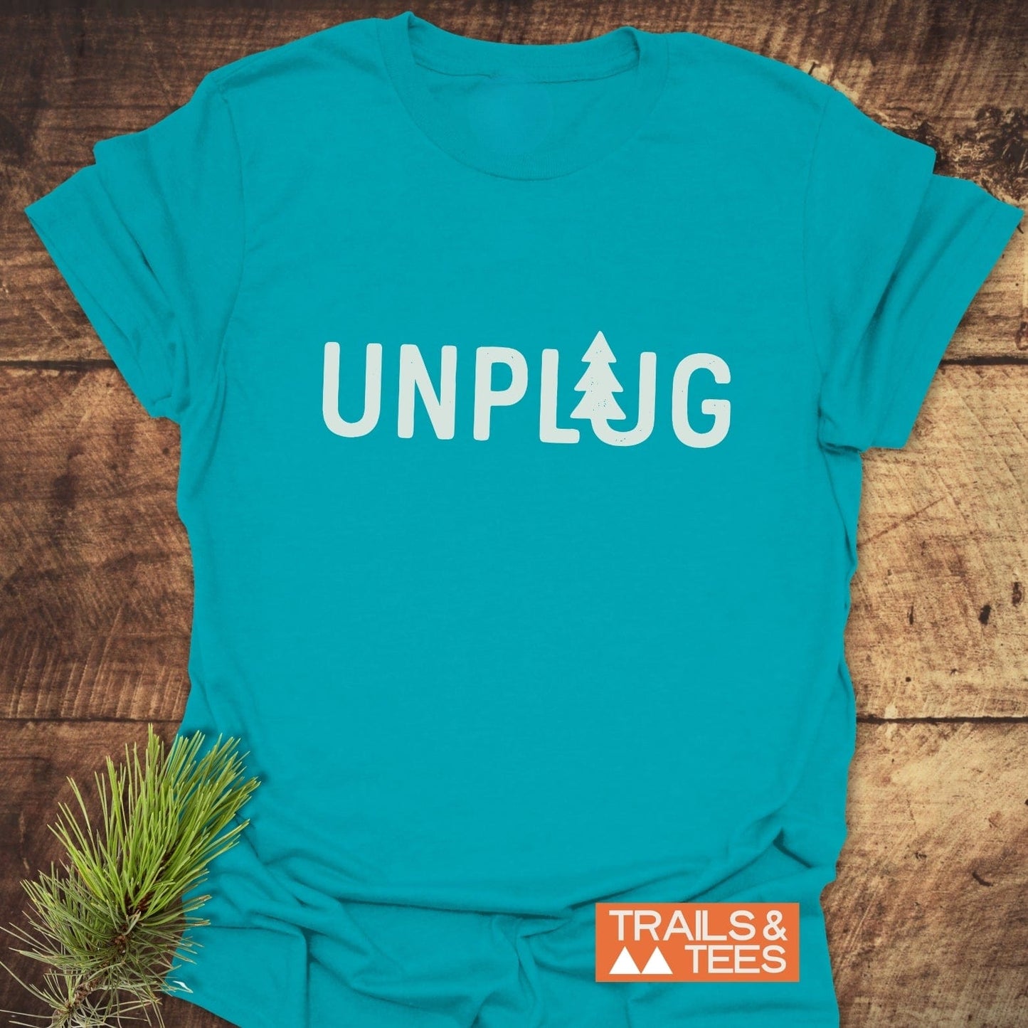 Unplug T-Shirt in teal with UNPLUG text and pine-tree icon, laid on wood. Features orange TRAILS & TEES logo, pine sprig accent.