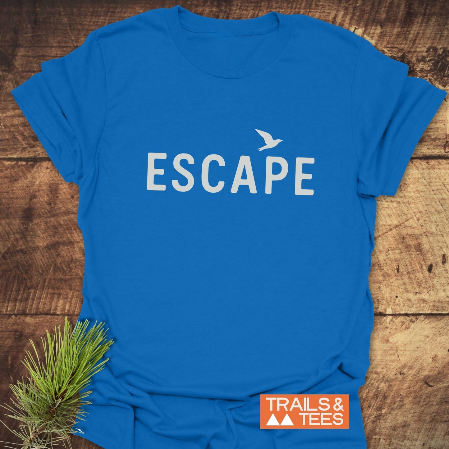 Escape Minimal T-Shirt featuring ESCAPE and bird design, laid on wood. Lightweight, ring-spun cotton for comfort, perfect for outdoor adventures.