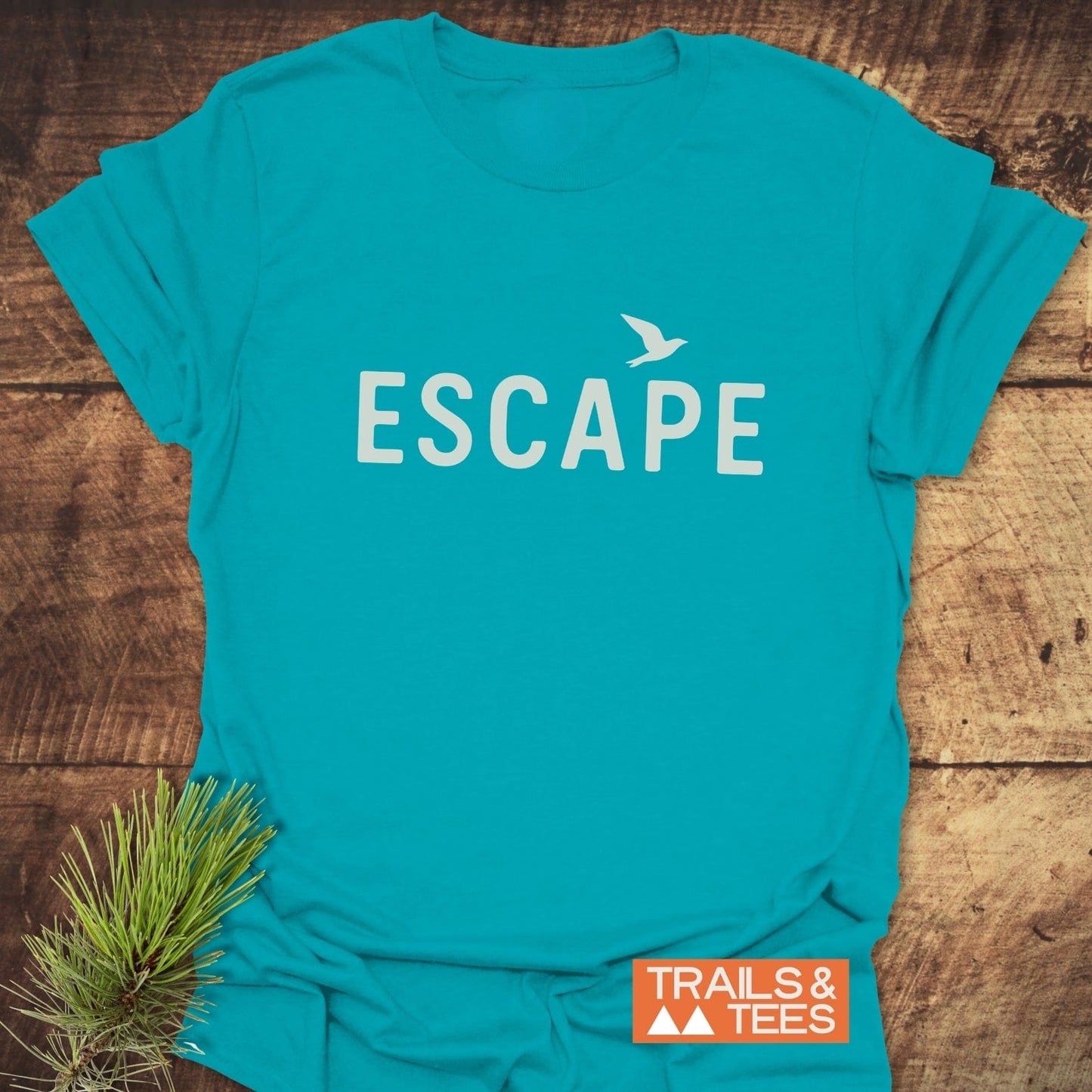 Escape Minimal T-Shirt featuring ESCAPE in white with a bird silhouette, laid on wood. Includes Trails & Tees logo, pine branch accent.