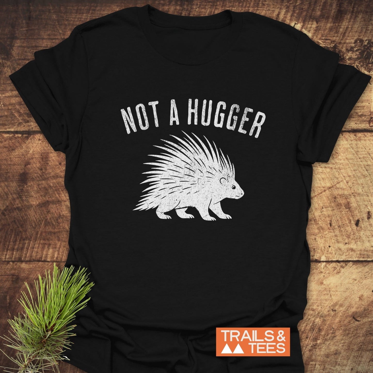 Not A Hugger T-Shirt featuring a distressed porcupine design, laid on wood with pine sprig. Soft cotton, durable print, pre-shrunk, logo in corner.