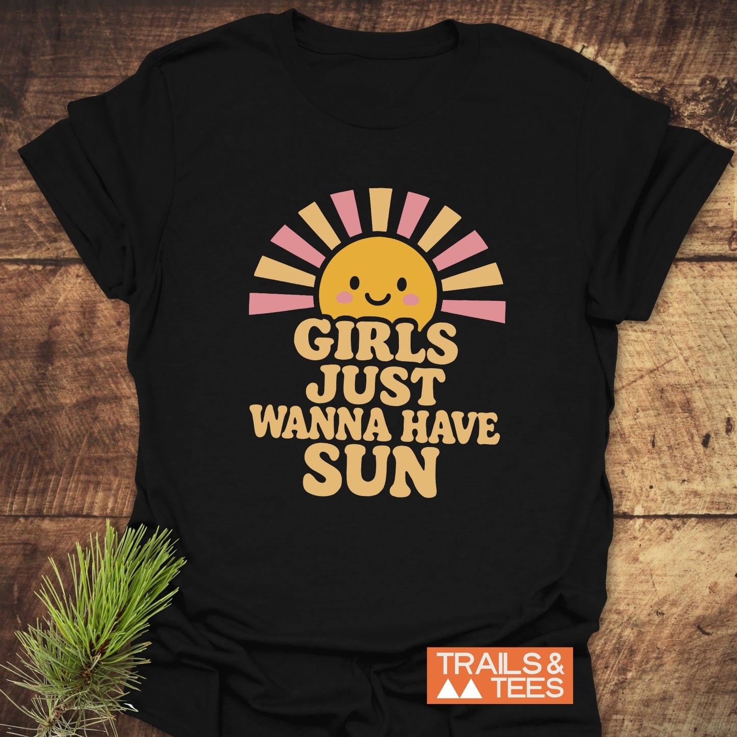 Girls Want Fun T-Shirt featuring a smiling sun with pink and golden rays, retro lettering, and the Trails & Tees logo on a wooden surface.