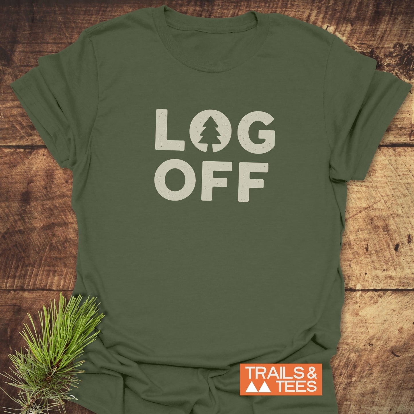Log Off T-Shirt featuring LOG OFF with a pine-tree logo on an olive-green base, laid flat with a nature-inspired pine sprig accentuating its outdoor theme.