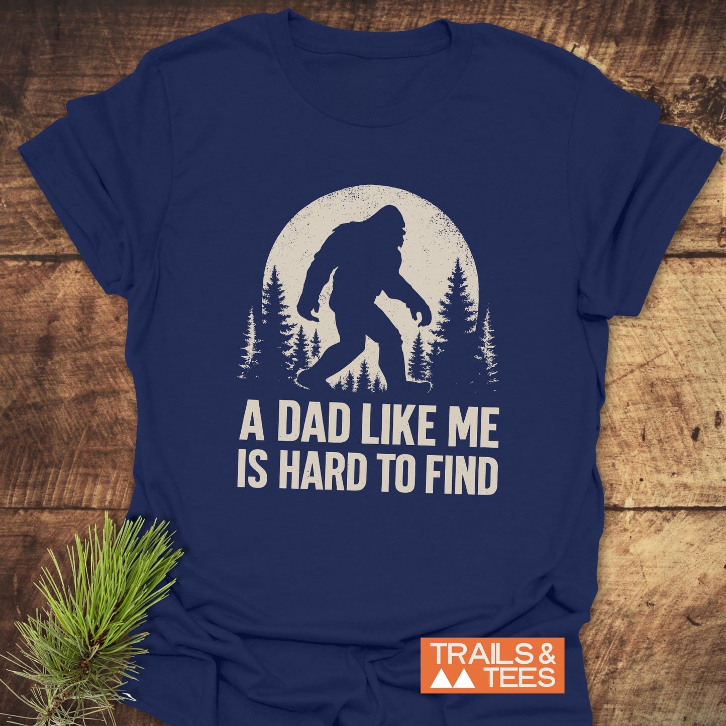 Vintage Bigfoot Dad T-Shirt on wooden surface, featuring a Bigfoot design with pine trees and moon, “A DAD LIKE ME IS HARD TO FIND” text.