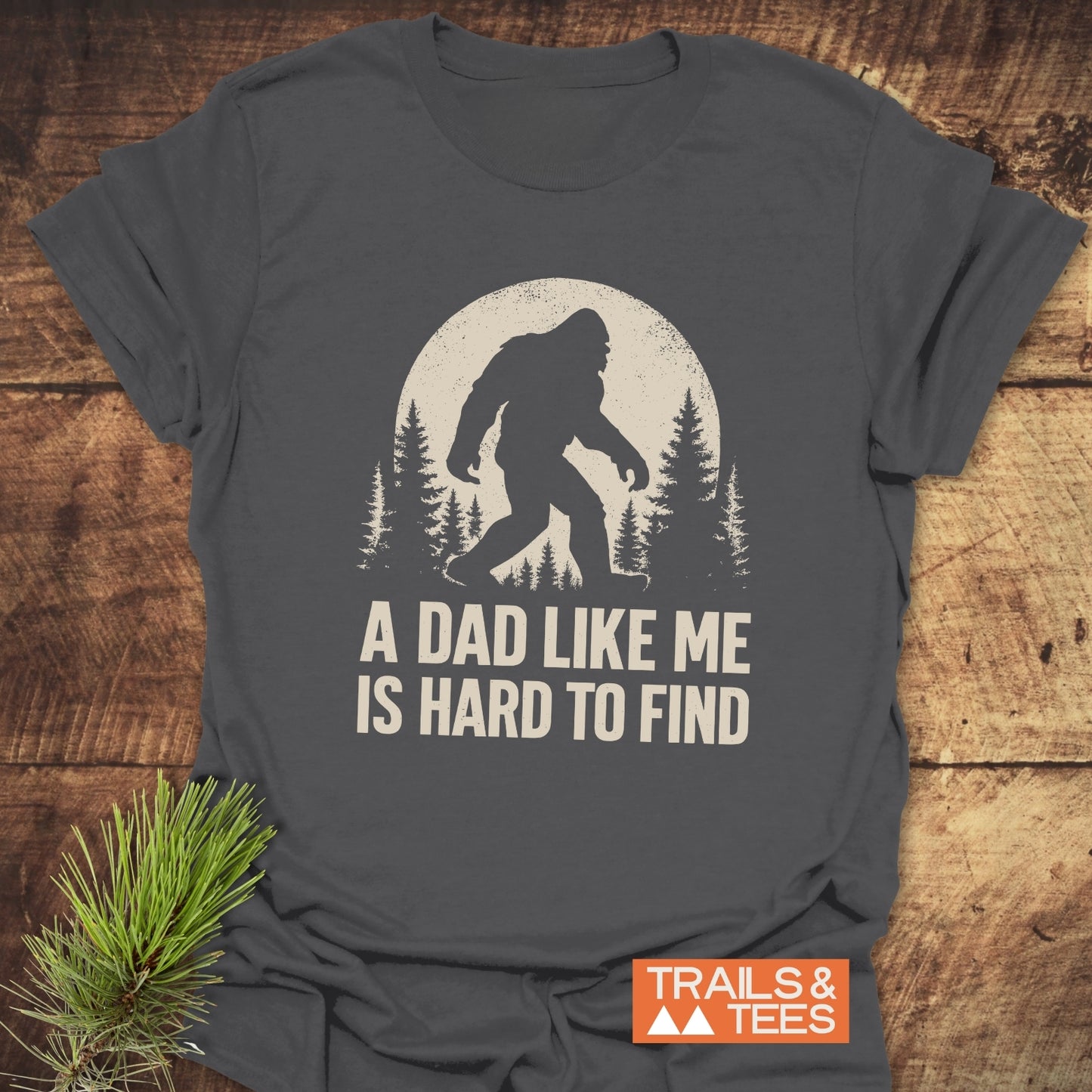 Vintage Bigfoot Dad T-Shirt with Sasquatch graphic and A DAD LIKE ME IS HARD TO FIND text, laid on wood, featuring durable, soft ring-spun cotton.