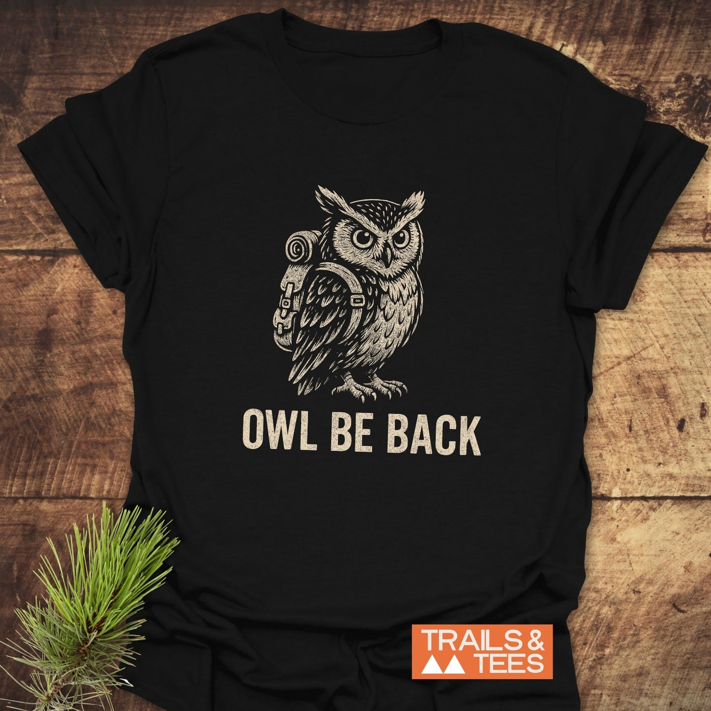 Owl Be Back Funny T-Shirt with an illustrated owl in a backpack and slogan, laid on wood. Durable, soft cotton for outdoor lovers, featuring Trails & Tees logo.