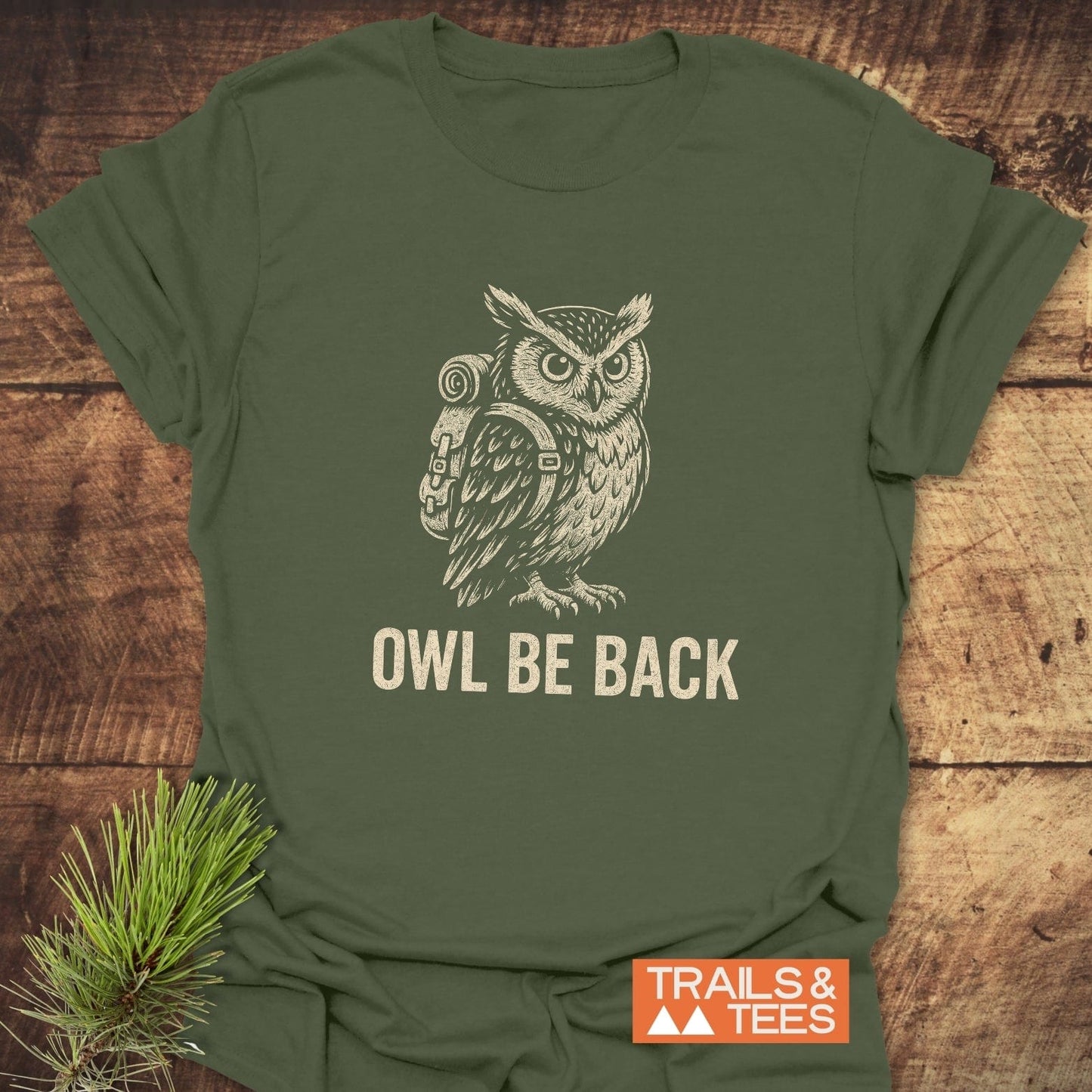Owl Be Back Funny T-Shirt featuring a hand-drawn owl with a backpack, slogan, and Trails & Tees logo on a wooden surface, accented with pine sprig.