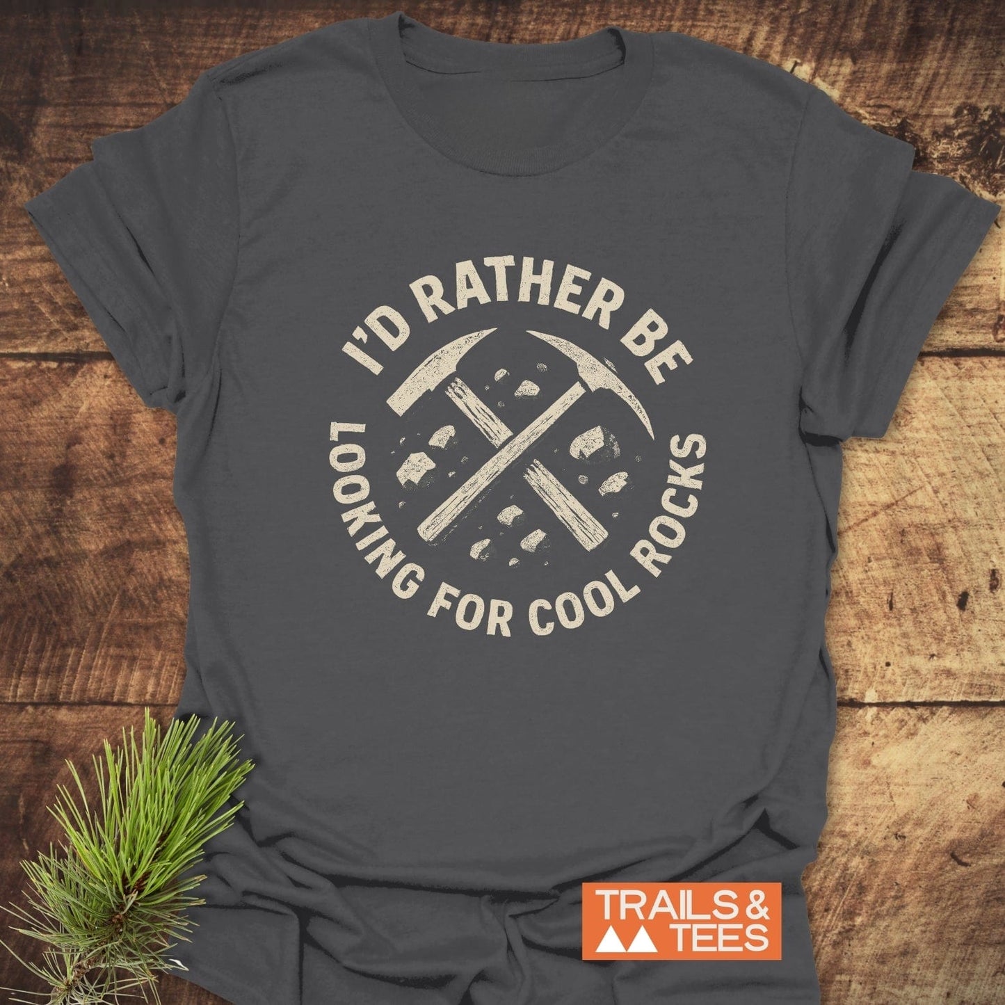 Dark-gray I'd Rather Be Looking for Cool Rocks T-shirt with rock-hammer graphic, laid on wood. Features durable print, soft cotton, no side seams.