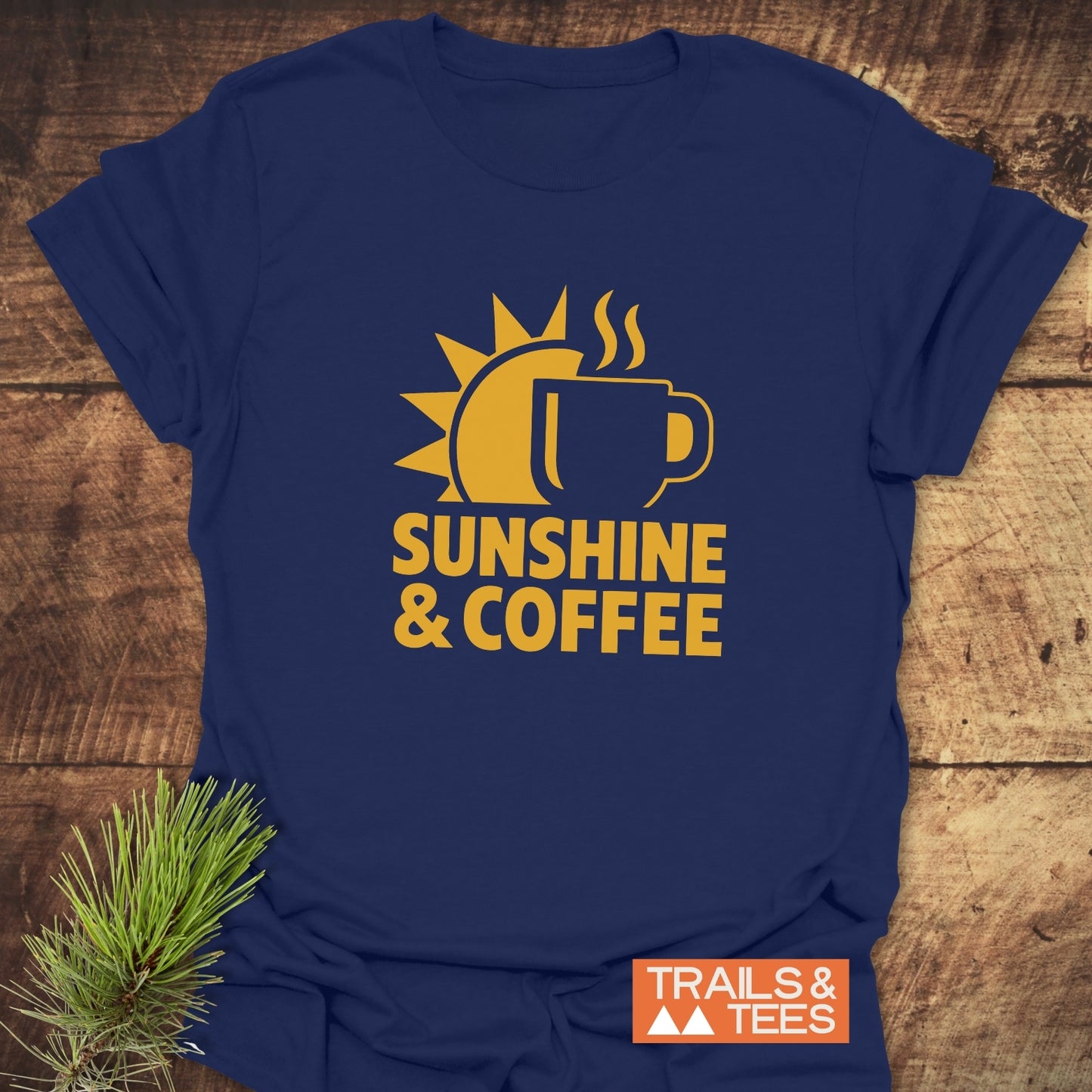 Bold Sunshine & Coffee T-Shirt with a half-sun and mug graphic, laid on wood, embodying adventure-ready comfort. Featuring durable print, pre-shrunk fit, and taped shoulders.