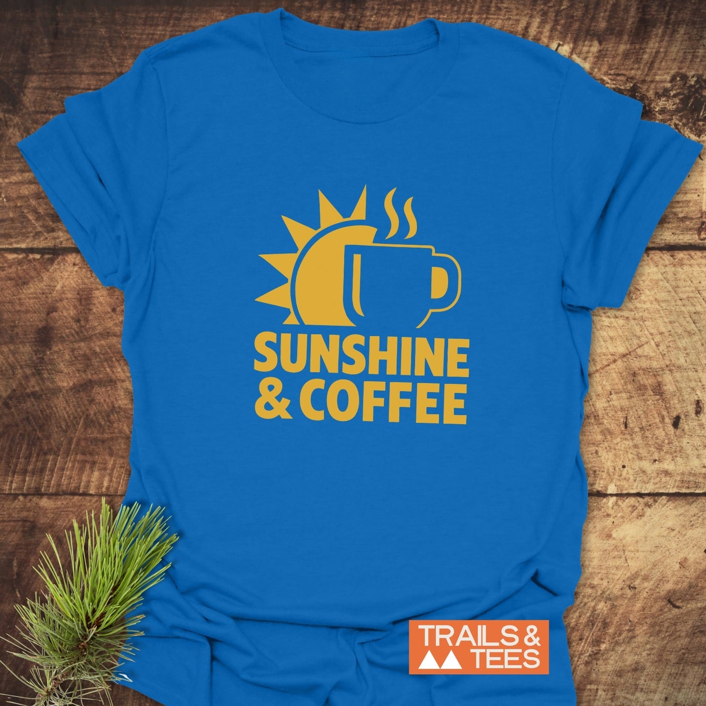 Bold Sunshine & Coffee T-Shirt featuring a sun and coffee mug graphic, laid flat with a pine sprig, embodies comfort and outdoor spirit by Trails & Tees.