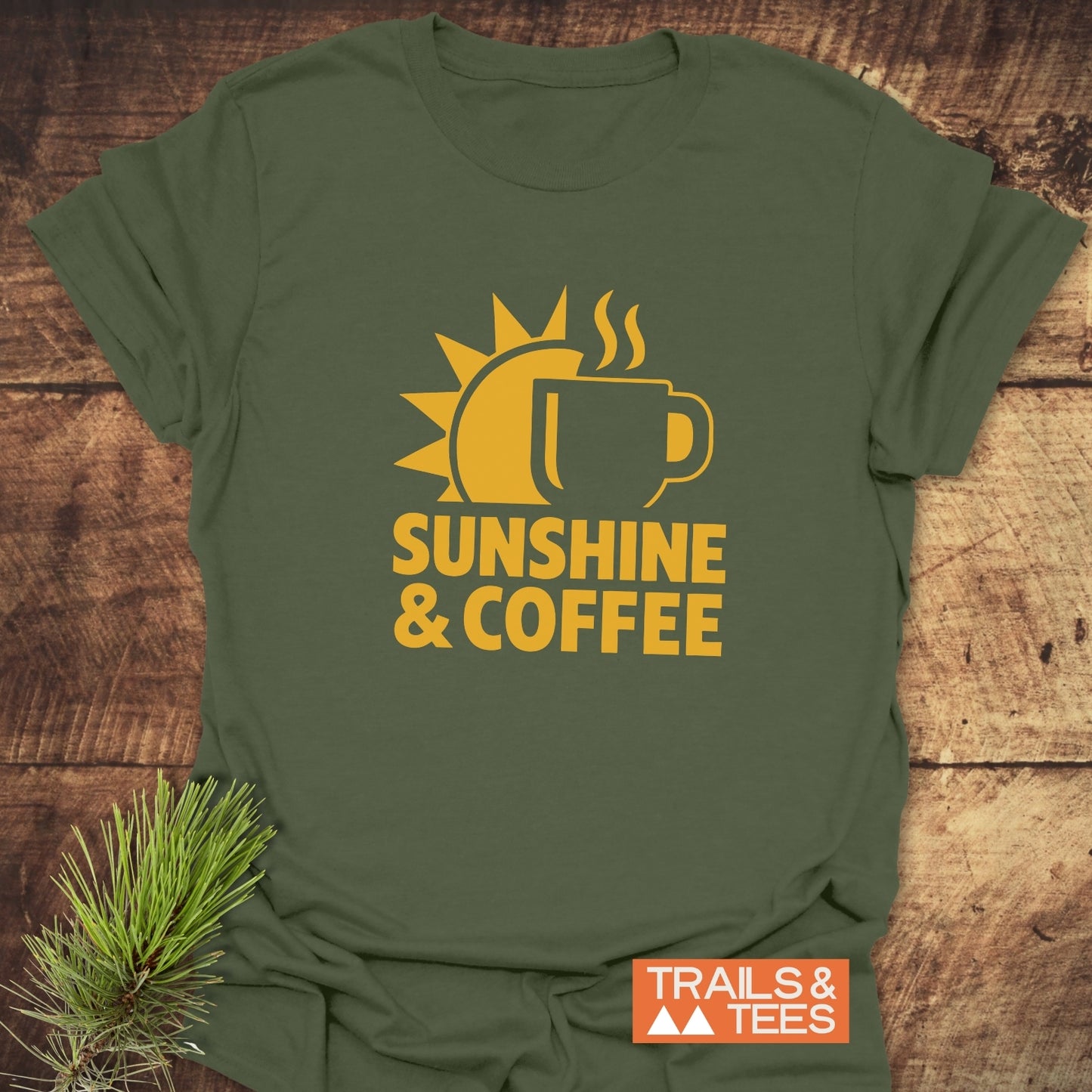 Bold Sunshine & Coffee T-Shirt featuring a rising sun and coffee mug graphic, laid flat with a TRAILS & TEES label on a wooden surface.