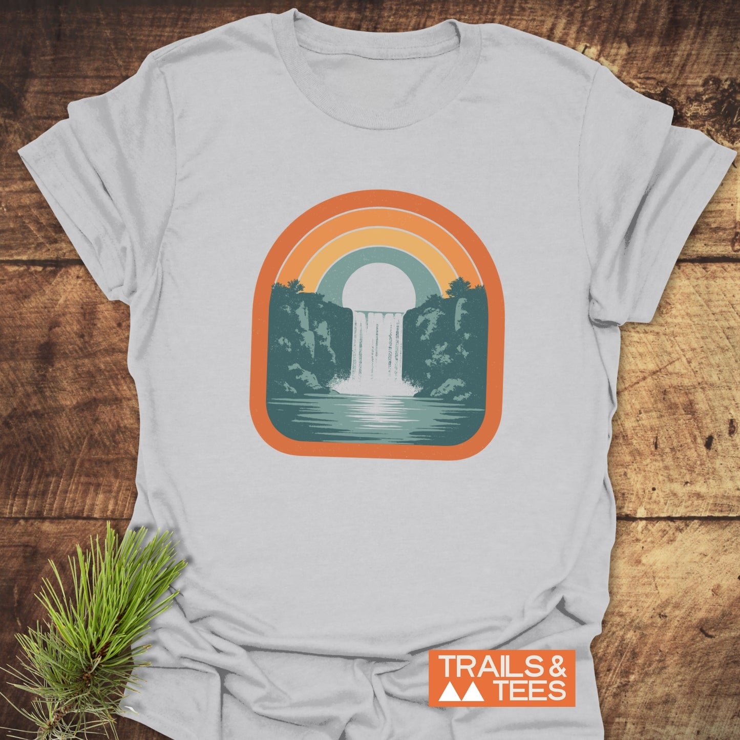 Vintage Waterfall T-Shirt featuring a retro waterfall graphic with orange, yellow, and teal arch, TRAILS & TEES branding, laid on wood with pine sprig.
