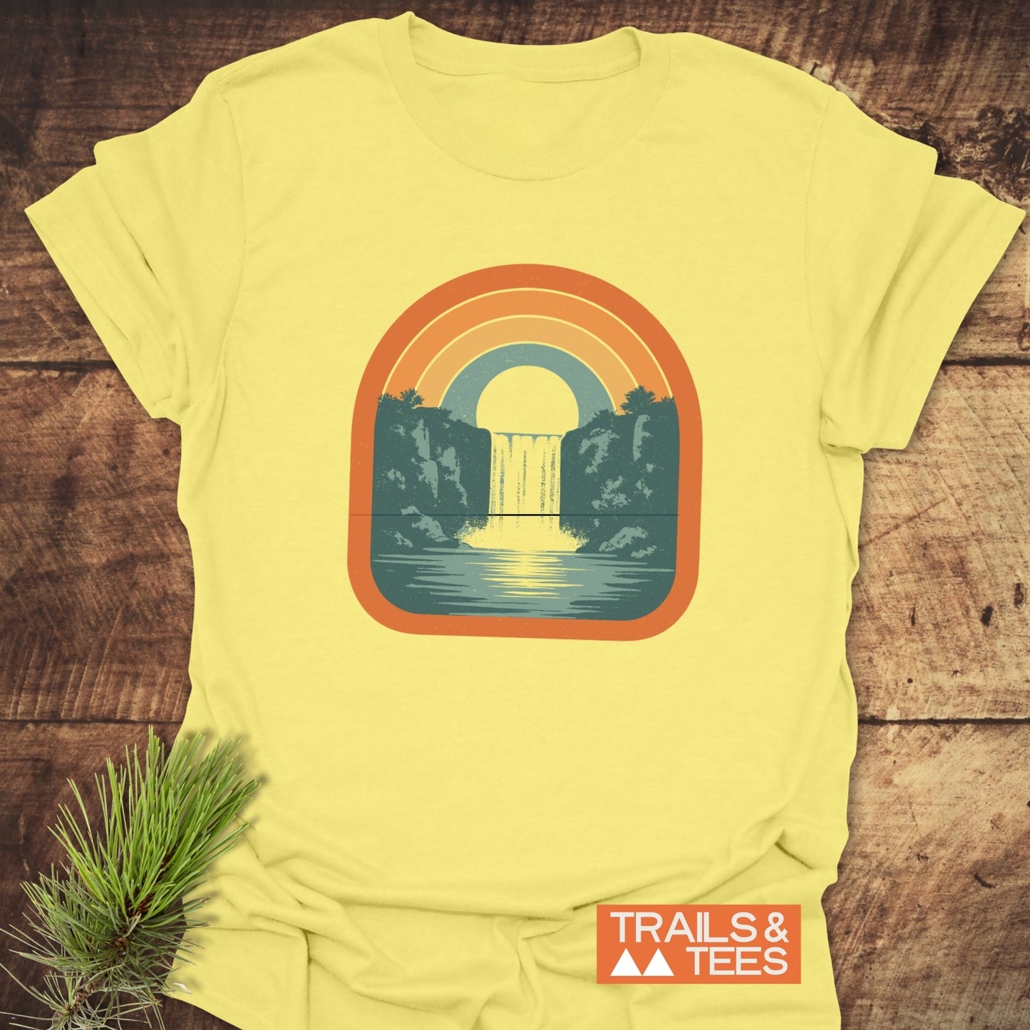 Vintage Waterfall T-Shirt featuring a retro waterfall graphic with sun and rainbow arches, laid on wood, highlighting outdoor-inspired, durable, pre-shrunk, ring-spun cotton design.