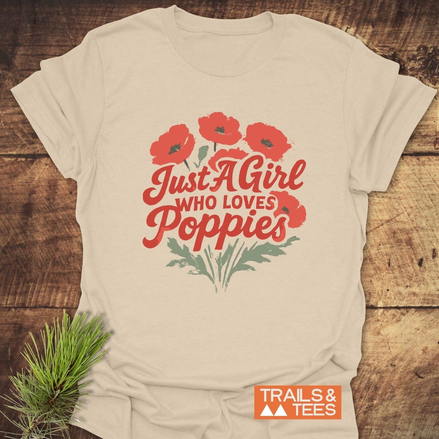 Girl Poppies T-Shirt with red poppy graphic and slogan on light-beige cotton, laid on wood. Includes TRAILS & TEES logo. Comfortable, durable, adventure-ready design.
