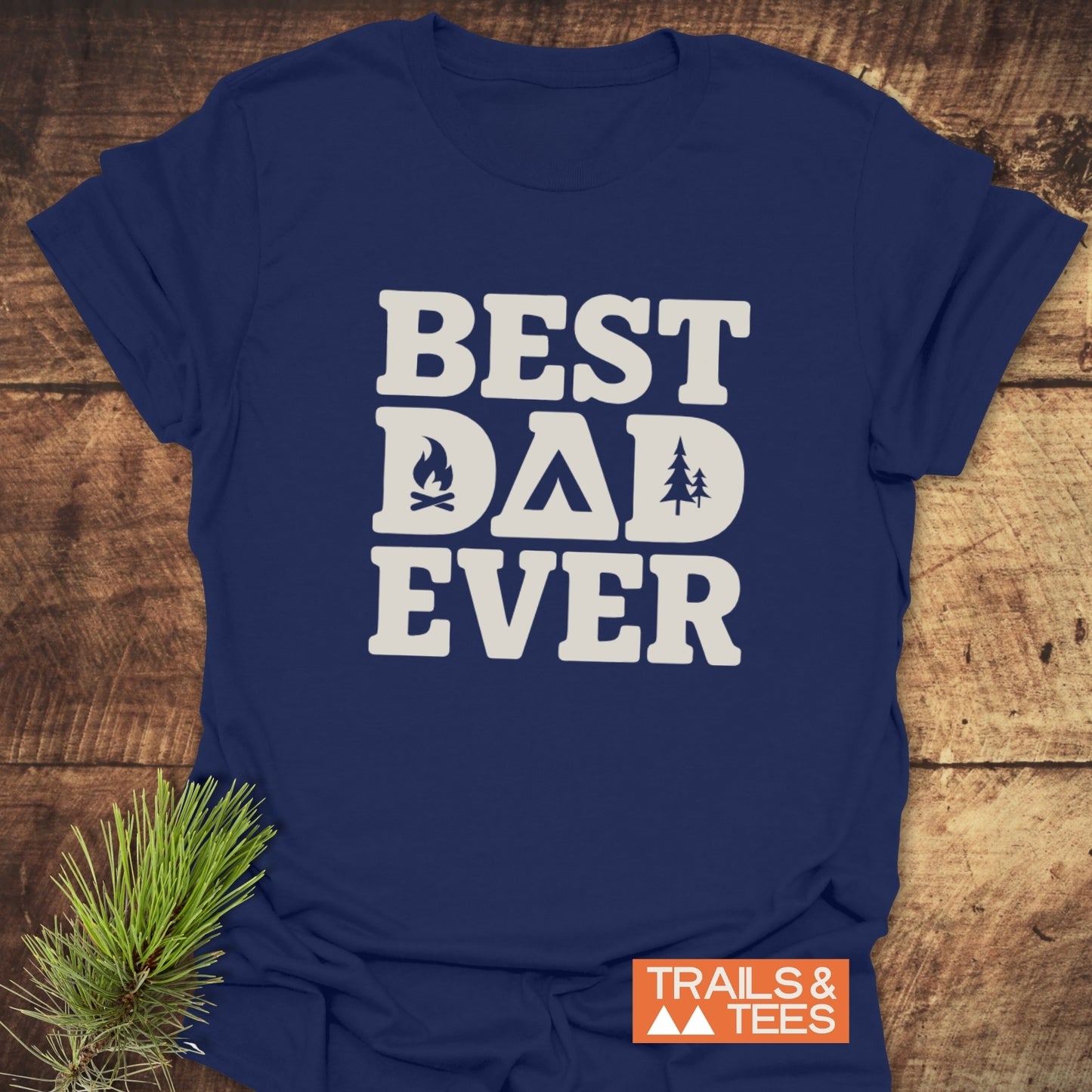 Navy-blue Best Dad Ever Camping T-Shirt with campfire, tent, and pine-tree icons, showcasing TRAILS & TEES logo, and a pine sprig nearby.