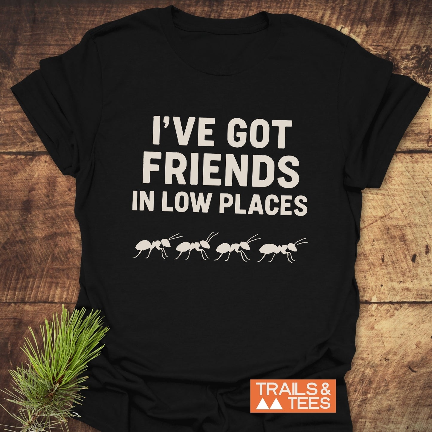 Friends In Low Places Funny T-Shirt featuring off-white text and ant silhouettes, crafted from soft cotton, with durable prints and a small orange Trails & Tees logo.