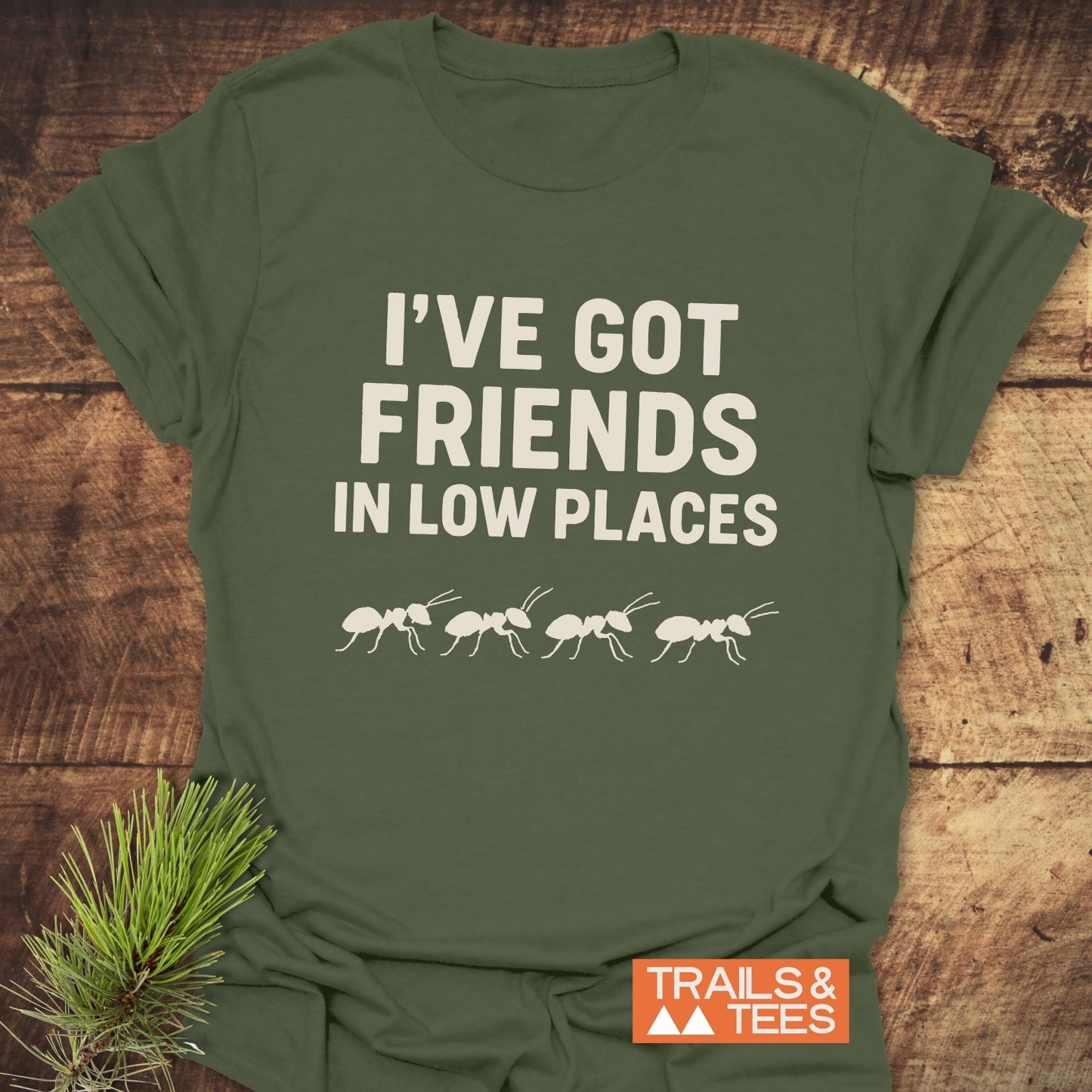 Friends In Low Places Funny T-Shirt featuring playful ant design, laid on wood. Made from soft cotton, perfect for outdoor enthusiasts.