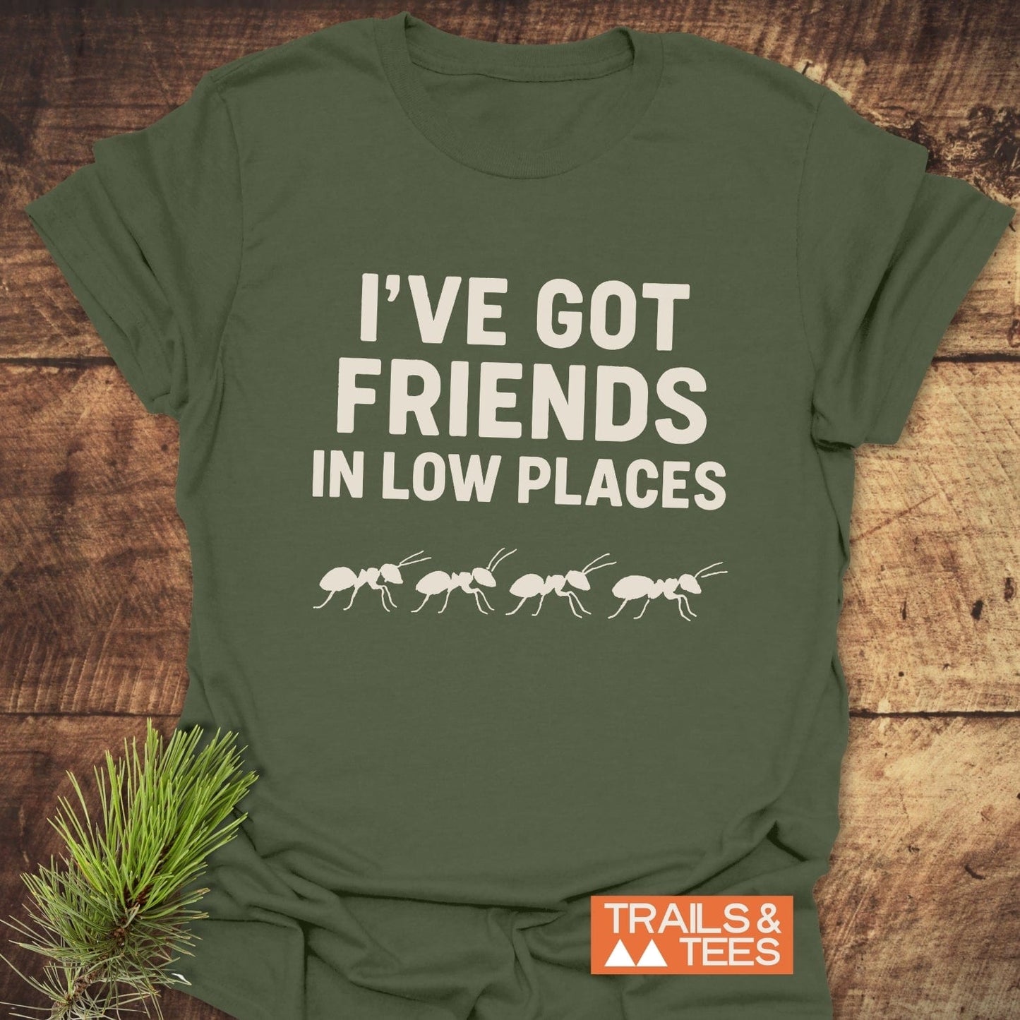 Friends In Low Places Funny T-Shirt featuring playful ant design, laid on wood. Made from soft cotton, perfect for outdoor enthusiasts.