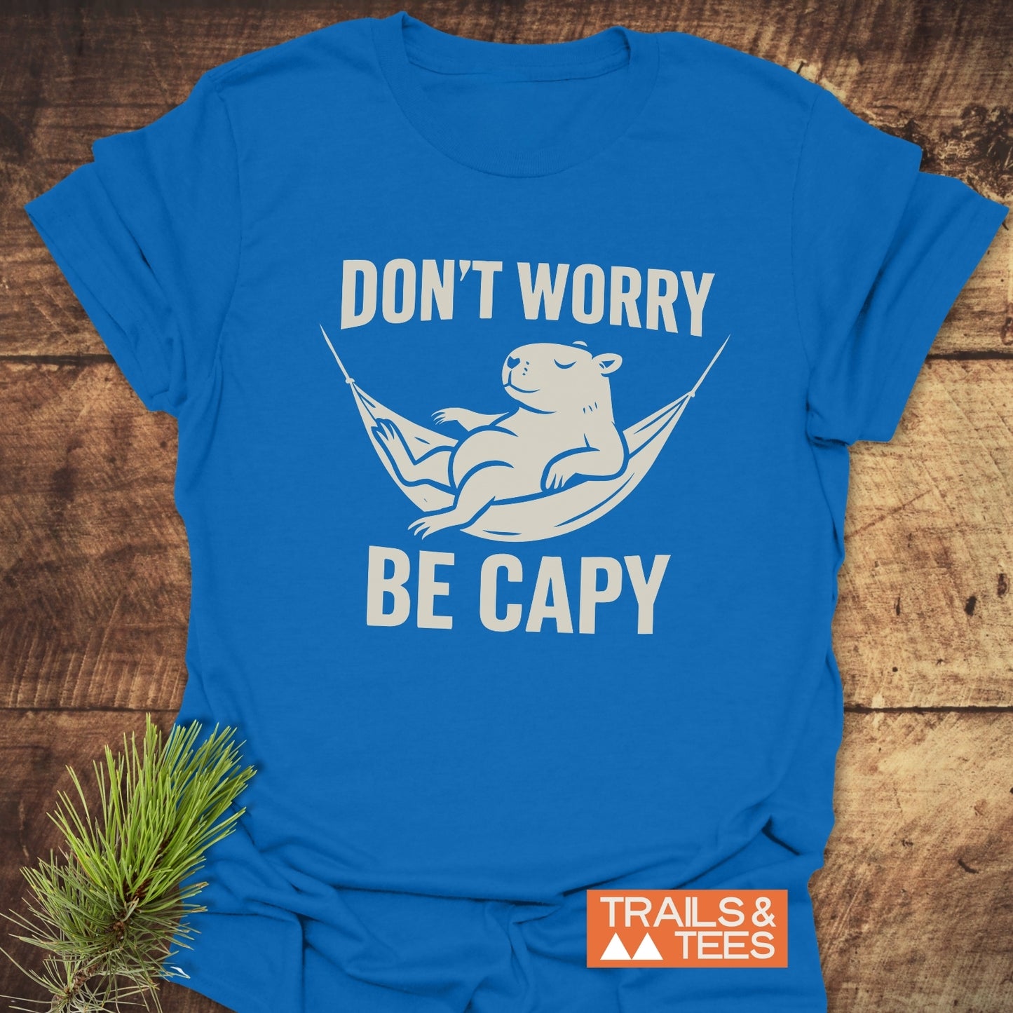 Don't Worry Capybara T-Shirt featuring a capybara in a hammock with DON’T WORRY BE CAPY slogan, Trails & Tees logo on wooden surface.