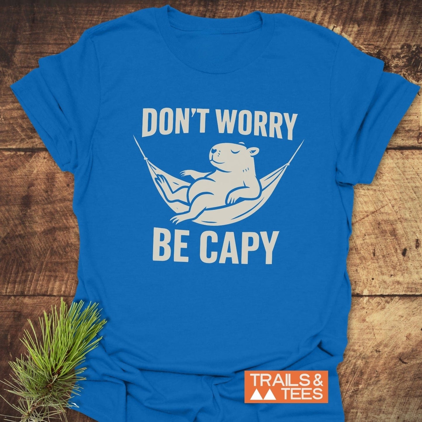 Don't Worry Capybara T-Shirt featuring a capybara in a hammock with DON’T WORRY BE CAPY slogan, Trails & Tees logo on wooden surface.