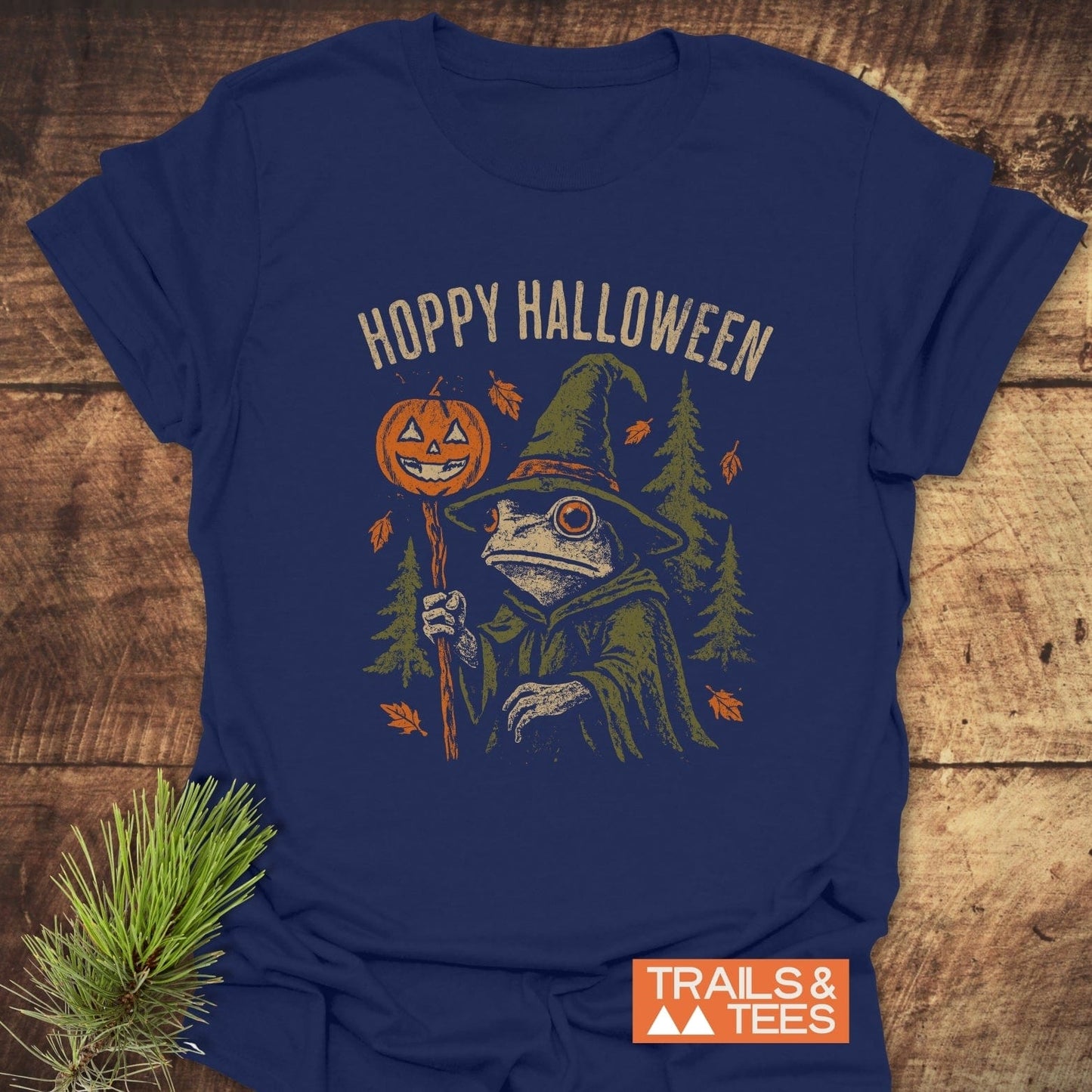 Hoppy Halloween T-Shirt featuring a witchy frog with jack-o’-lantern staff amidst autumn leaves and pine trees, highlighting Trails & Tees' outdoor-inspired design.