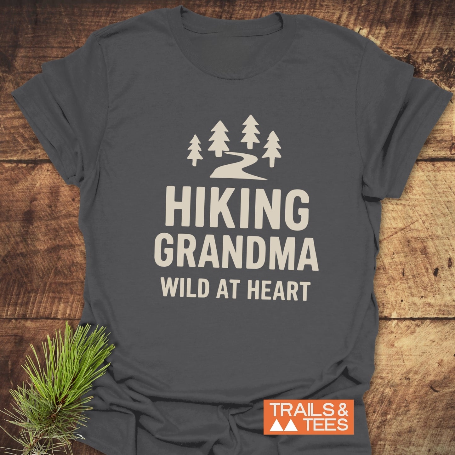 Dark gray Hiking Grandma - Wild At Heart T-shirt with pine tree graphic, laid on wood. Features soft cotton, durable print, taped shoulders.