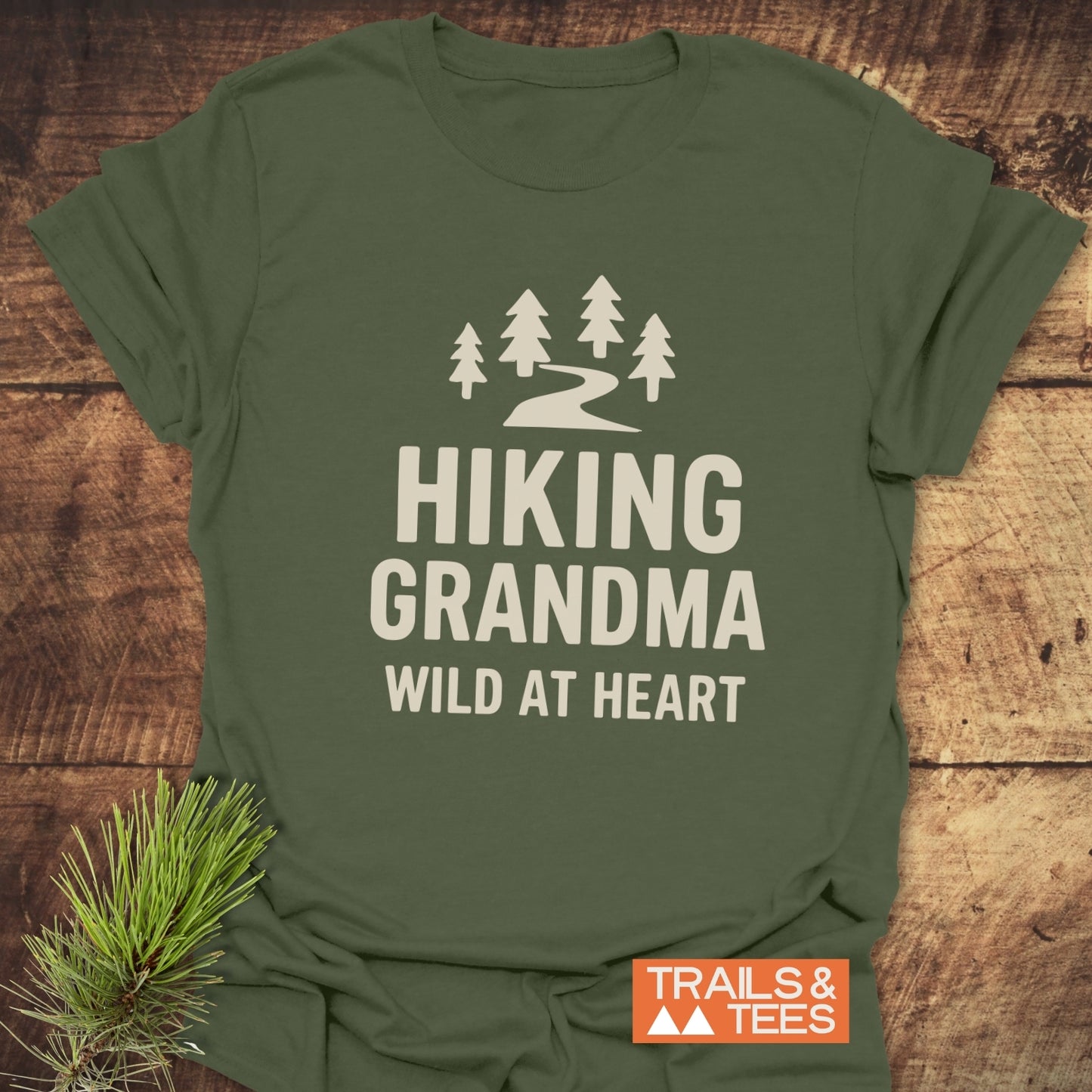 Olive-green tee with pine trees and Hiking Grandma Wild At Heart text, highlighting outdoor adventure spirit. Includes brand TRAILS & TEES and evergreen sprig accent.