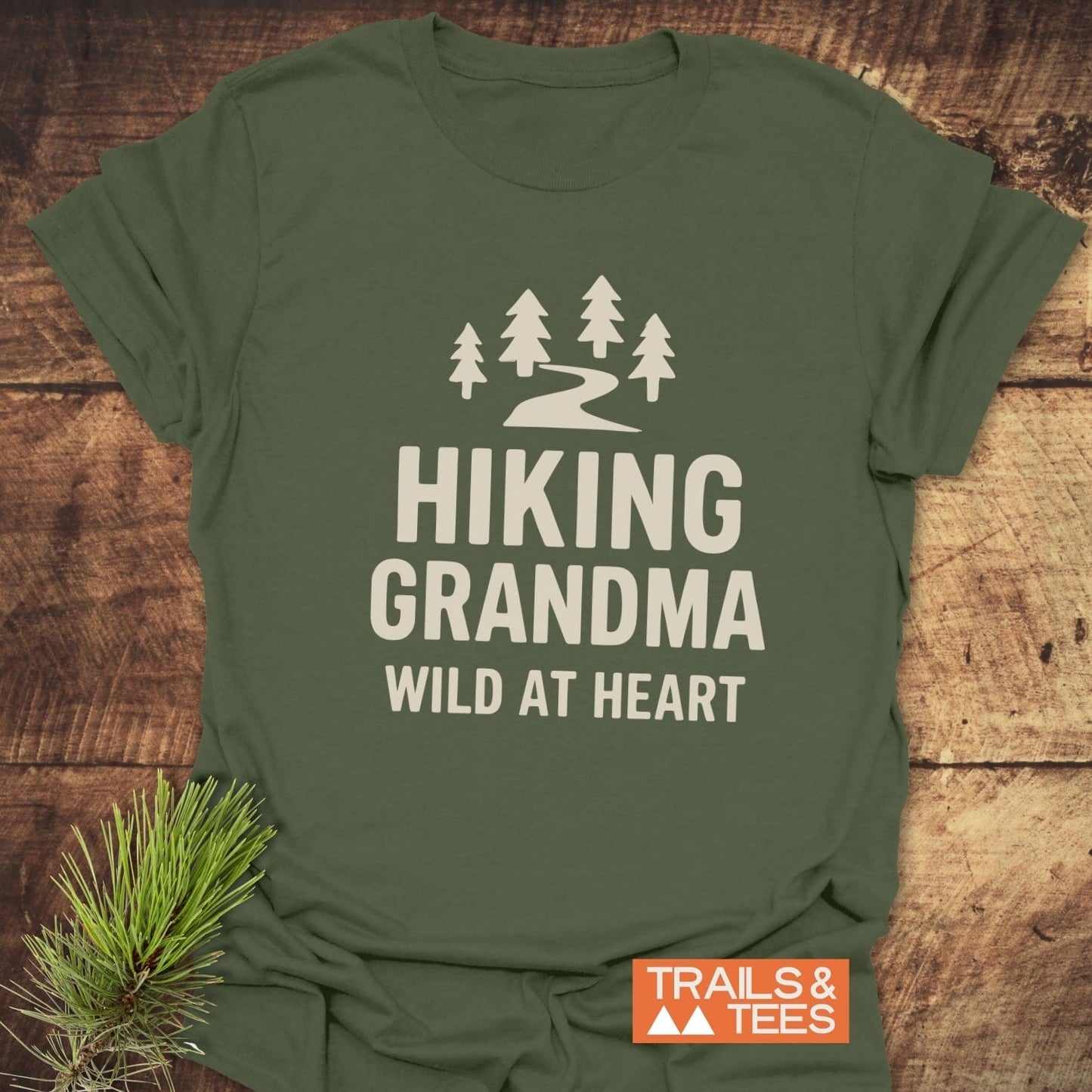 Olive-green tee with pine trees and Hiking Grandma Wild At Heart text, highlighting outdoor adventure spirit. Includes brand TRAILS & TEES and evergreen sprig accent.