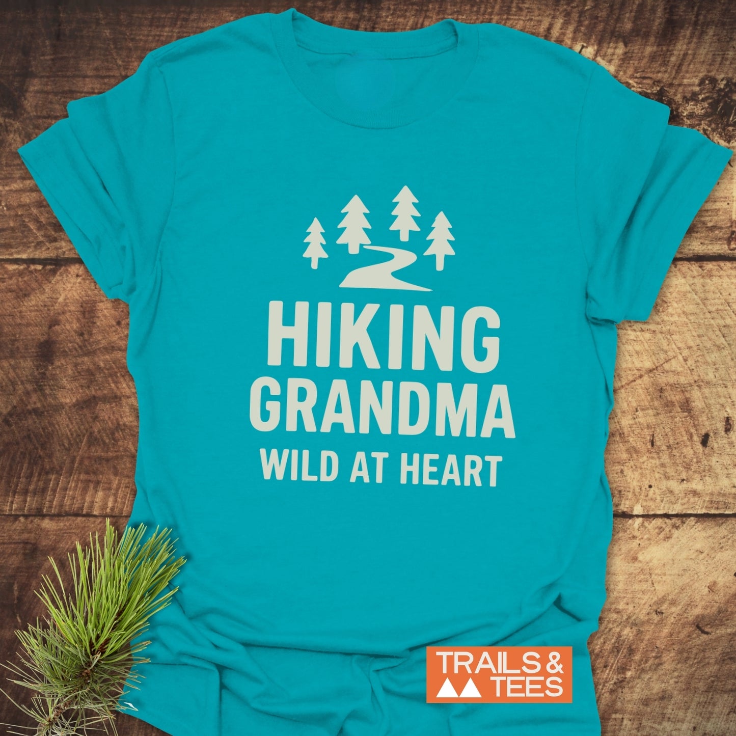 Hiking Grandma - Wild At Heart T-Shirt featuring a white trail and pine graphic, laid flat with pine sprig, showcasing outdoor-inspired comfort and durable design.