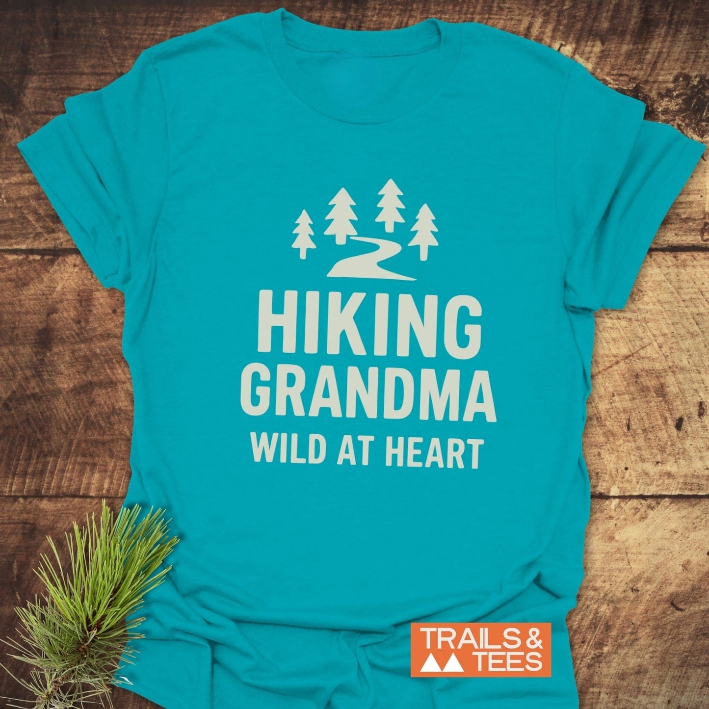 Hiking Grandma - Wild At Heart T-Shirt featuring a white trail and pine graphic, laid flat with pine sprig, showcasing outdoor-inspired comfort and durable design.
