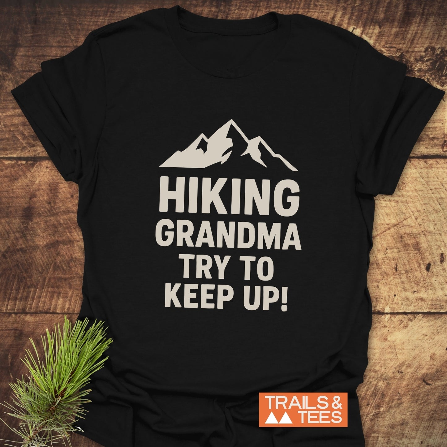 Black “Hiking Grandma - Try To Keep Up” T-shirt with mountain silhouette on wooden surface, featuring durable print and soft cotton for outdoor adventures.