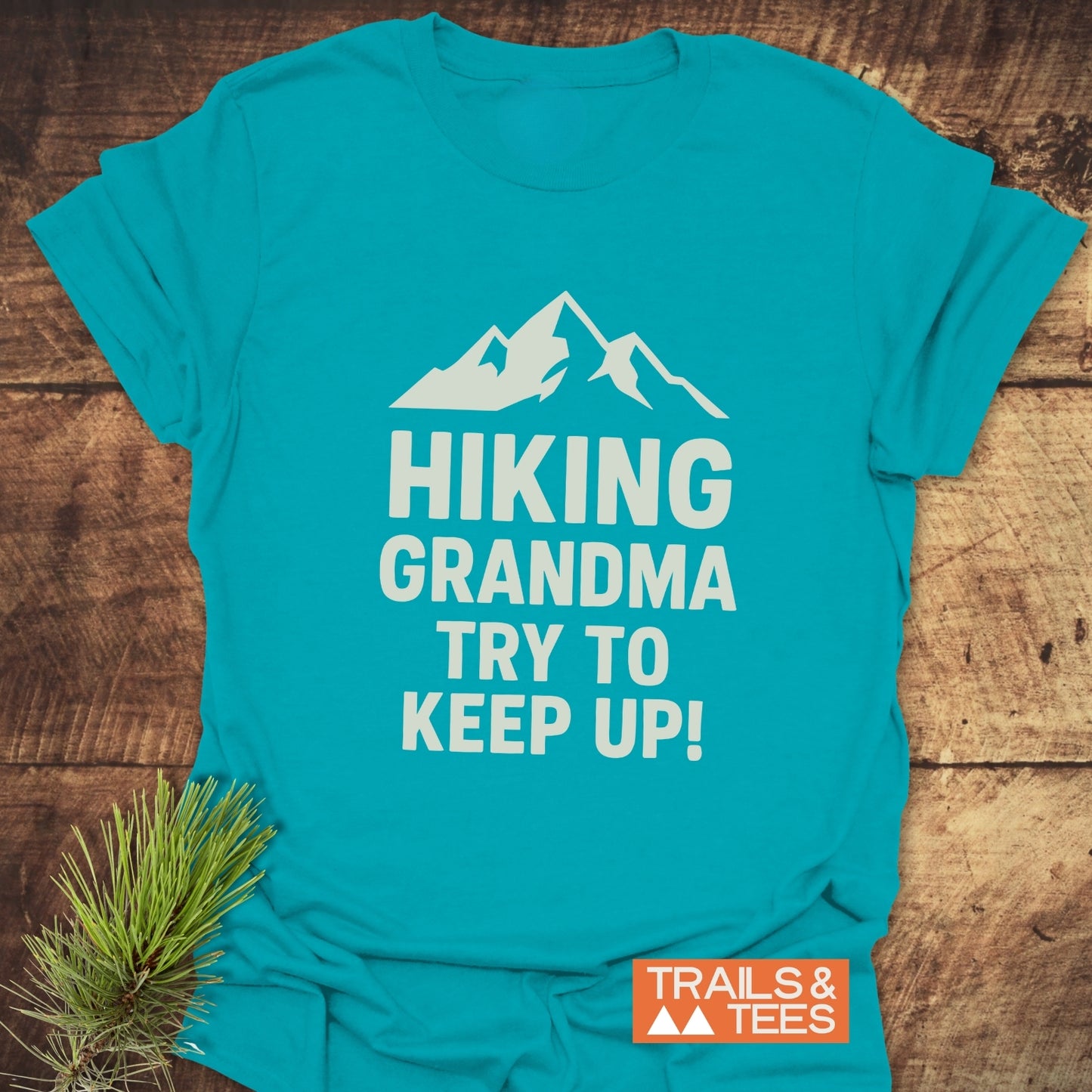 Hiking Grandma - Try To Keep Up T-Shirt, featuring a mountain silhouette and bold text, placed on a wooden surface with pine sprig for rustic charm.